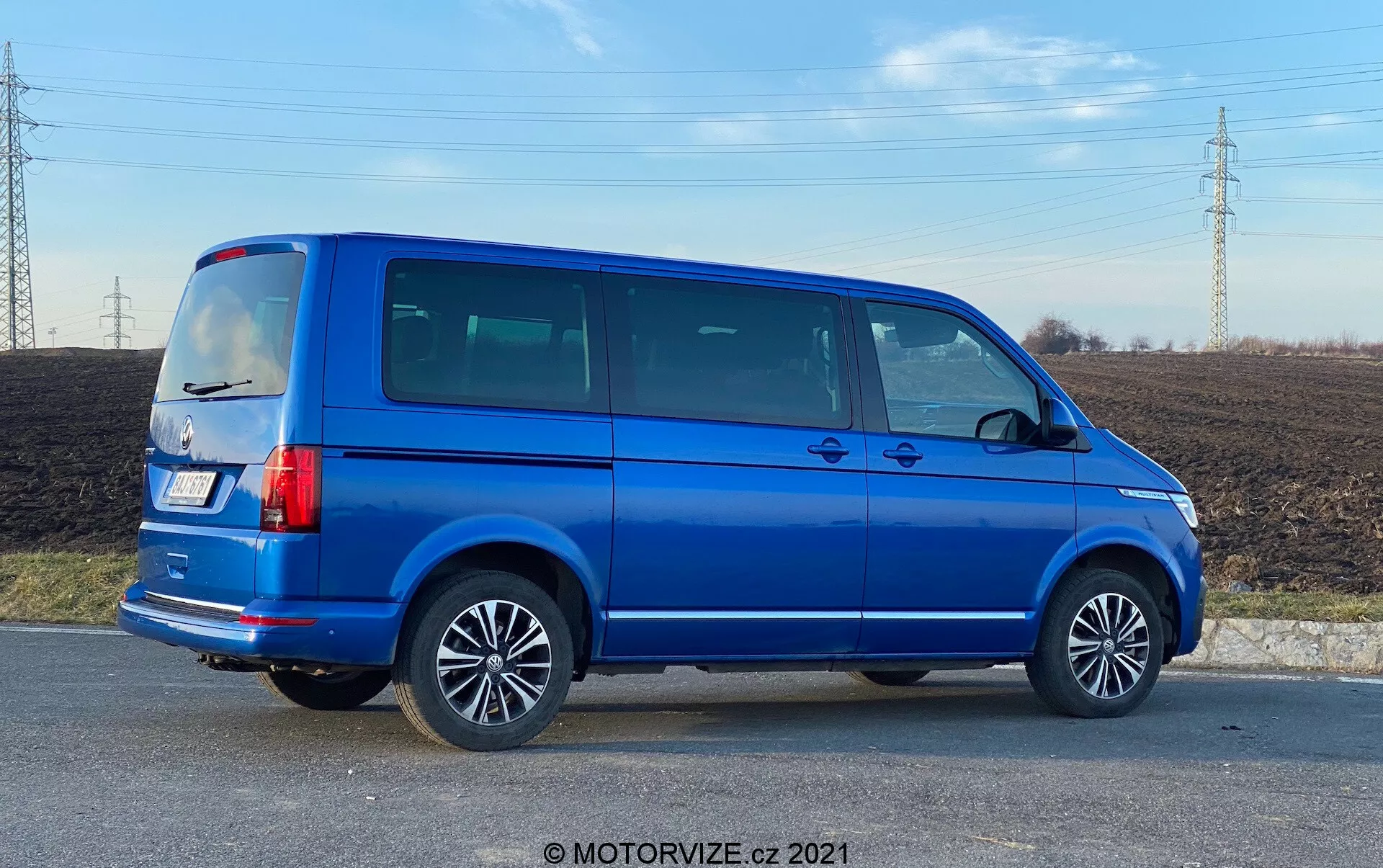 Kompletný bočný pohľad na modrý 2019 VW Nutzfahrzeuge Transporter (T6.1 Multivan) s hliníkovými kolesami, viditeľnými líniami dverí, tónovanými bočnými oknami, hranatou siluetou, vertikálnymi zadnými svetlami, anténou na streche a jasne modrým kovovým lakom. Obrázok je nasnímaný pod uhlom, čím poskytuje perspektívu na bočné a zadné časti vozidla, nachádzajúce sa vo vidieckej oblasti s poľom a elektrickými vedeniami v pozadí, za denného svetla.