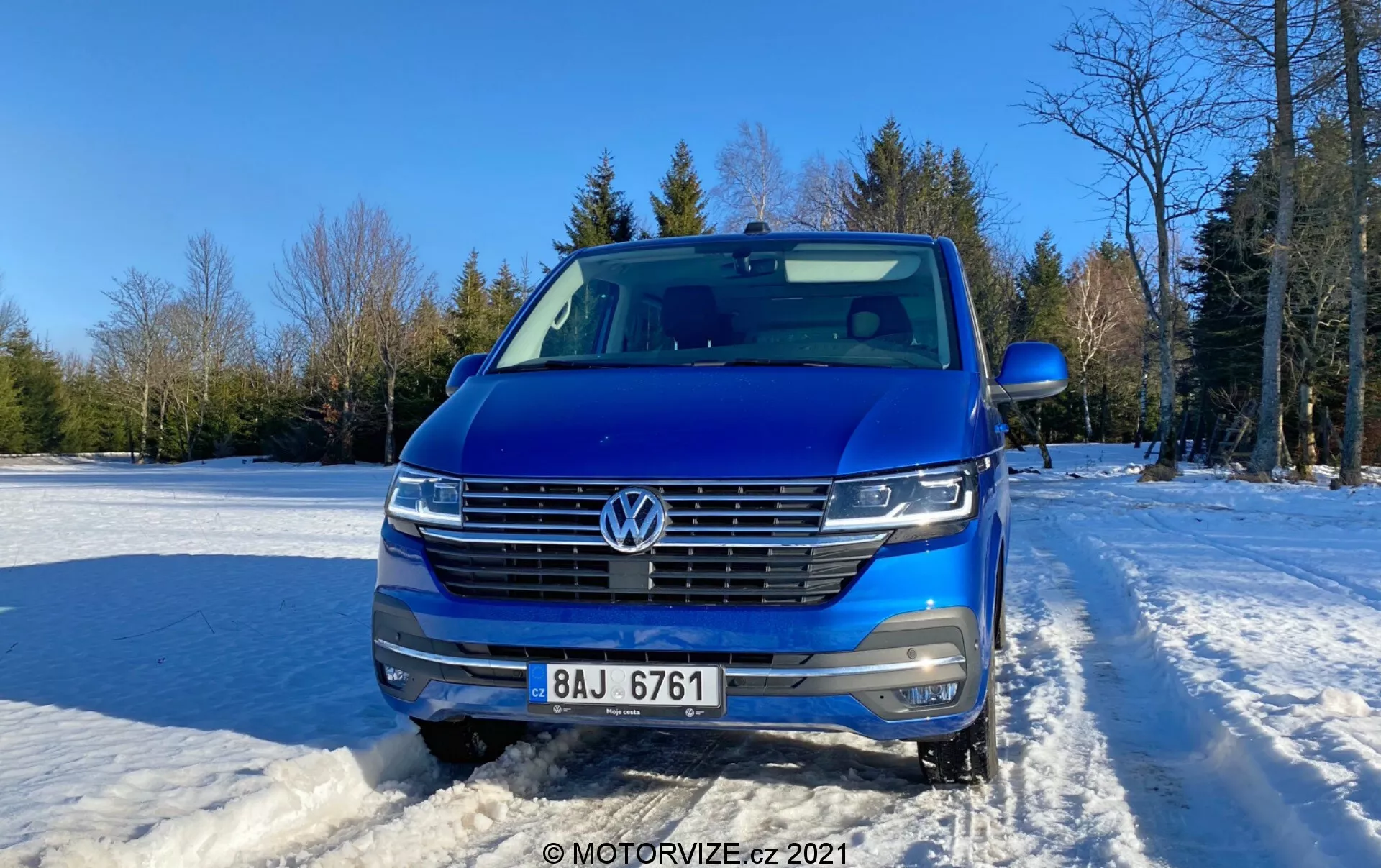 Čelný záber na modrý Volkswagen Transporter (2019) (T6-1 Multivan), ktorý ukazuje mriežku, svetlomety a kapotu. Vozidlo je umiestnené vonku na zasneženom povrchu s jasnou oblohou a stromami v pozadí. Vlastnosti auta zahŕňajú emblém VW, horizontálnu mriežku so zrkadlovým lemom, svetlomety LED, kameru smerujúcu dopredu, parkovacie senzory, strieborné strešné lišty a čiastočne viditeľné bočné zrkadlo. Moderný dizajn s aerodynamickým predkom, vyfotografovaný cez deň s jemným slnečným svetlom z pravej strany.