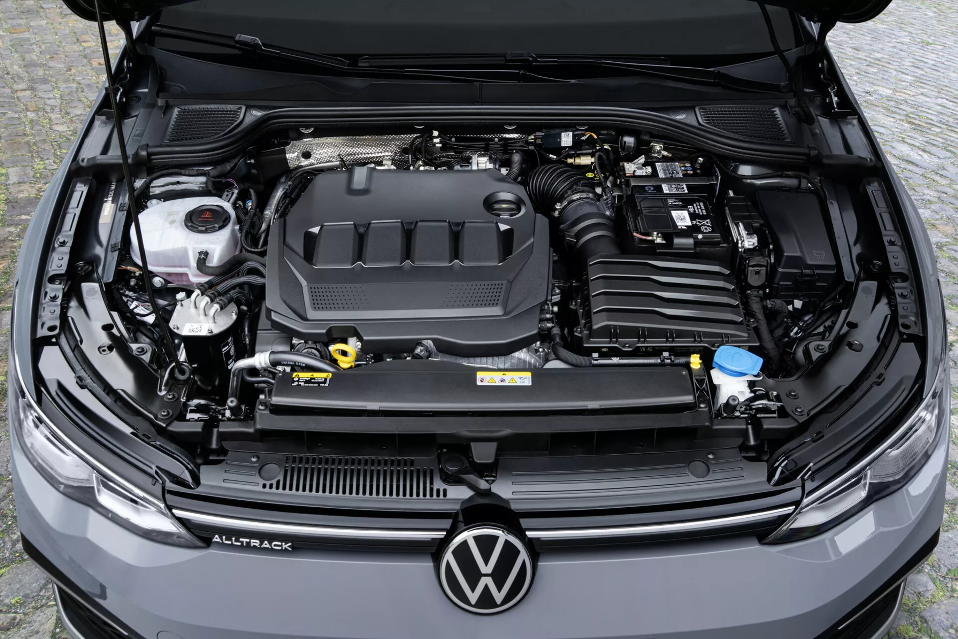 Pohľad zhora na detailný pohľad na motorový priestor Volkswagen Golf Alltrack 2017 s viditeľným krytom motora, zásobníkom na brzdovú kvapalinu, nádržkami na chladiacu kvapalinu a kvapalinu ostrekovačov, spolu s servisnými bodmi a etiketami s informáciami o vozidle, ktoré predvádzajú moderné funkcie automobilovej technológie a stav údržby vozidla.