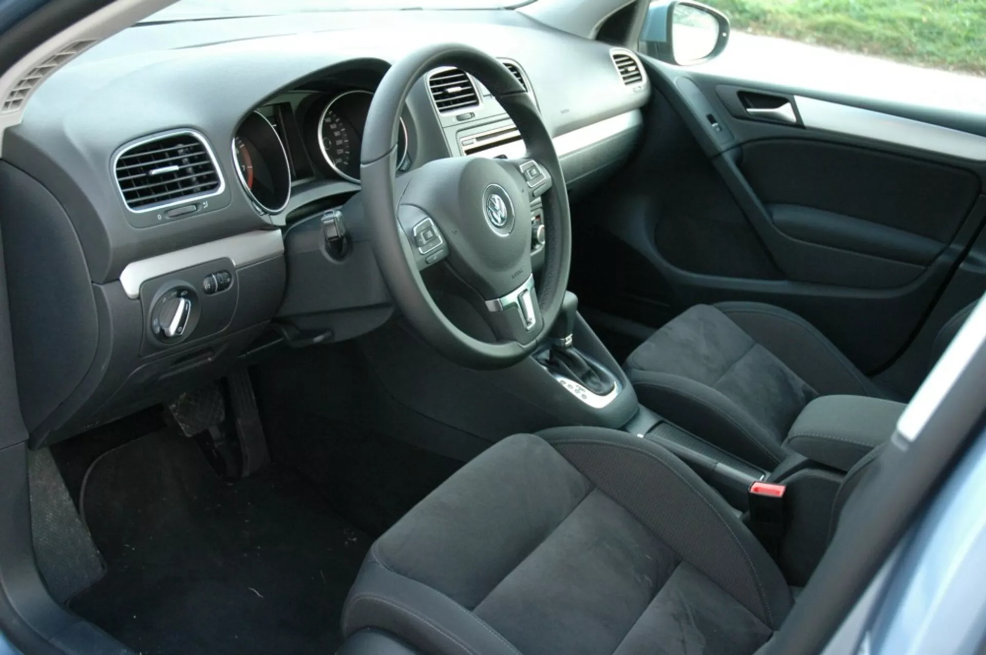 Interiér VW Golf 2008 zobrazujúci stranu vodiča z pohľadu zadného pasažiera s volantom, prístrojovou doskou, stredovou konzolou, radiacou pákou automatickej prevodovky, sedadlami potiahnutými tmavou látkou, ovládacími prvkami na dverách vodiča, kobercovou podlahou s rohožami a prirodzeným osvetlením