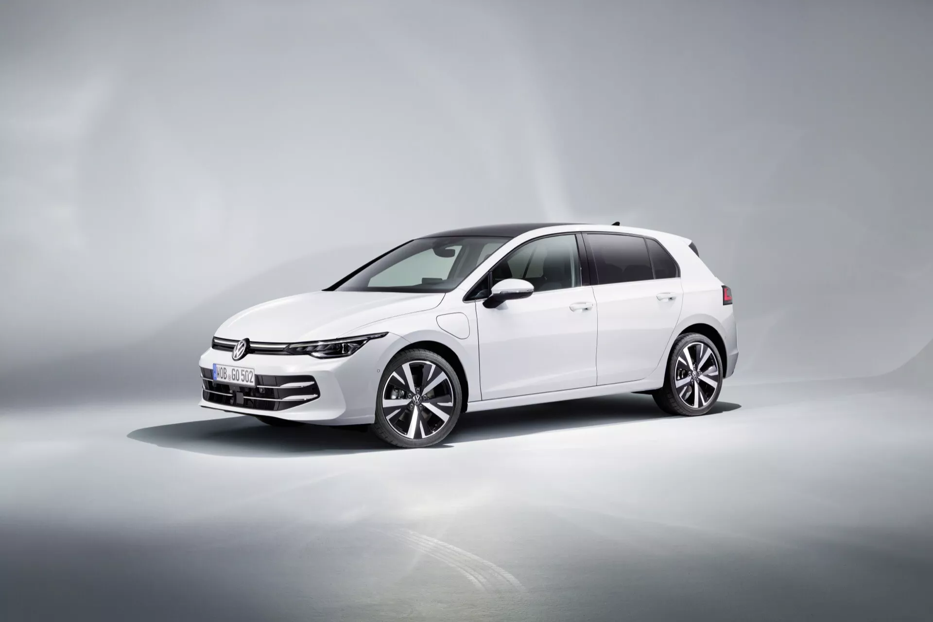 VW Golf (VIII-Facelift) 2024 v bielej farbe, zobrazuje dynamický trojštvrťový predný pohľad na odrazivej povrchove so sfarbeným pozadím, zdôrazňujúci jeho úzke LED svetlomety, vodorovnú mriežku, viacramenné zliatinové kolesá, zrkadlá so zabudovanými smerovkami a tónované okná.