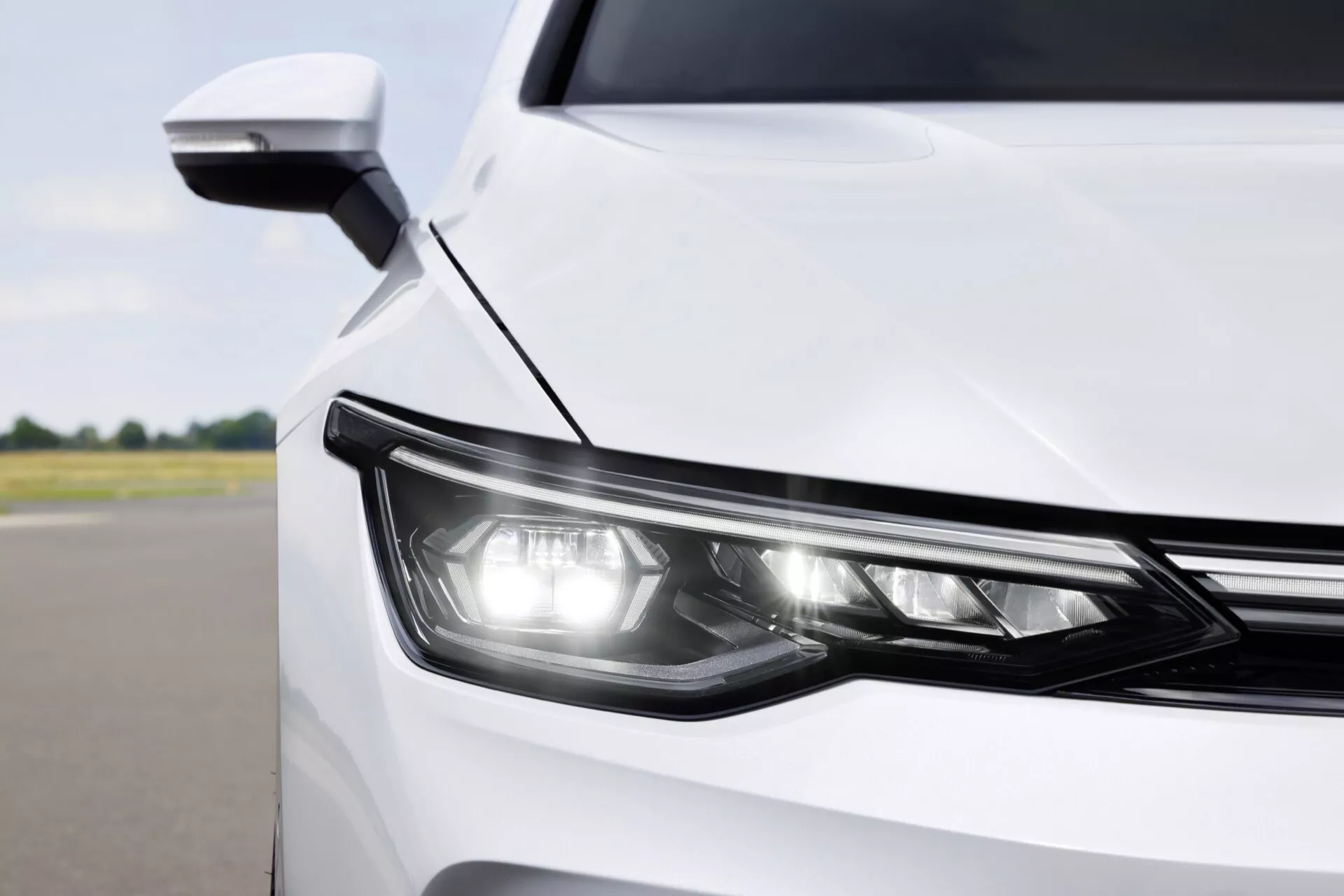 Detailný pohľad na predný roh bieleho VW Golf VIII facelift 2024, ukazujúci moderný dizajn a technológiu LED svetlometu a aerodynamického bočného zrkadla s integrovaným smerovým ukazovateľom, z čiastočného pohľadu z mierne zvýšeného uhla