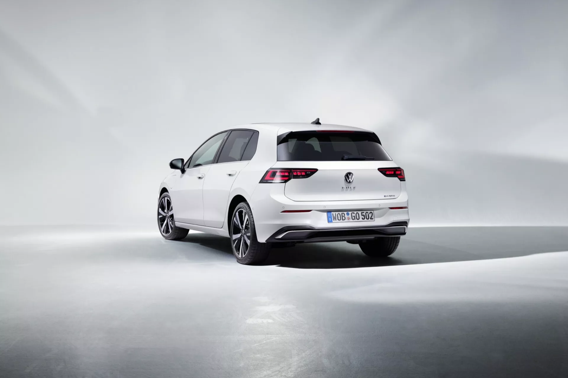 Facelift Volkswagen Golf VIII 2024 v bielom vyhotovení predvádza LED zadné svetlá, športový zadný nárazník s integrovanou výfukovou špicou, dizajn hatchbacku, kolesá z ľahkých zliatin vysokej kvality a potenciálnu technológiu asistencie pri parkovaní, všetko zachytené z dynamického trojštvrťového zadného uhlu s mäkkým osvetlením, ktoré zdôrazňuje dizajnové krivky na jednoduchom pozadí.
