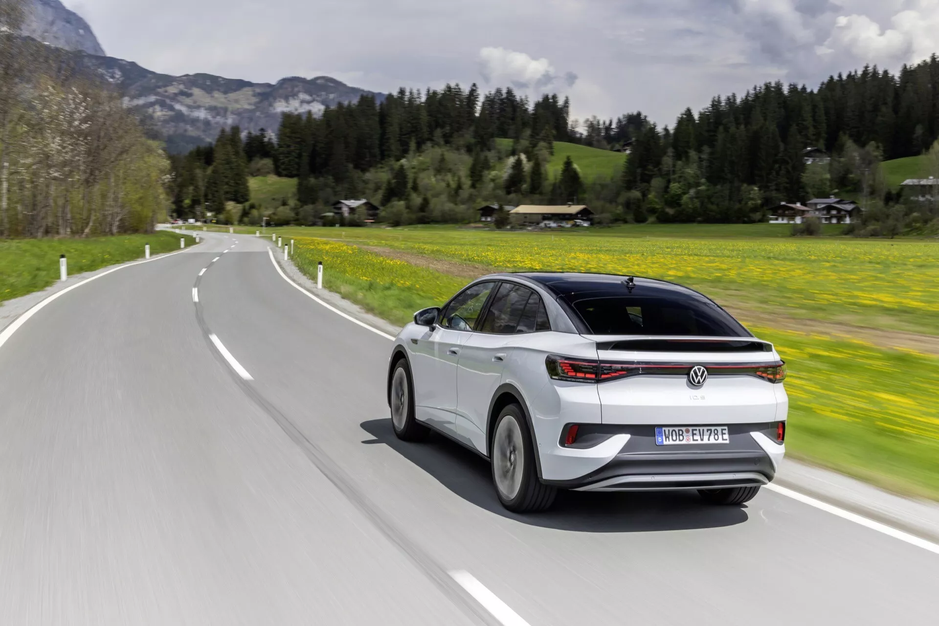 Dynamický trojštvrťový pohľad zozadu na pohybujúci sa VW ID.5 2022 (1. generácia) na ceste. Uhol zdôrazňuje elegantný zadný dizajn vozidla a bočný profil, ukazuje zadné prvky ako kufor, zadné sklo, zadné svetlá a zadný nárazník spolu s technologickými prvkami ako LED zadné svetlá a bočné zrkadlá s integrovanými smerovkami na pozadí bujnej zelenej krajiny.