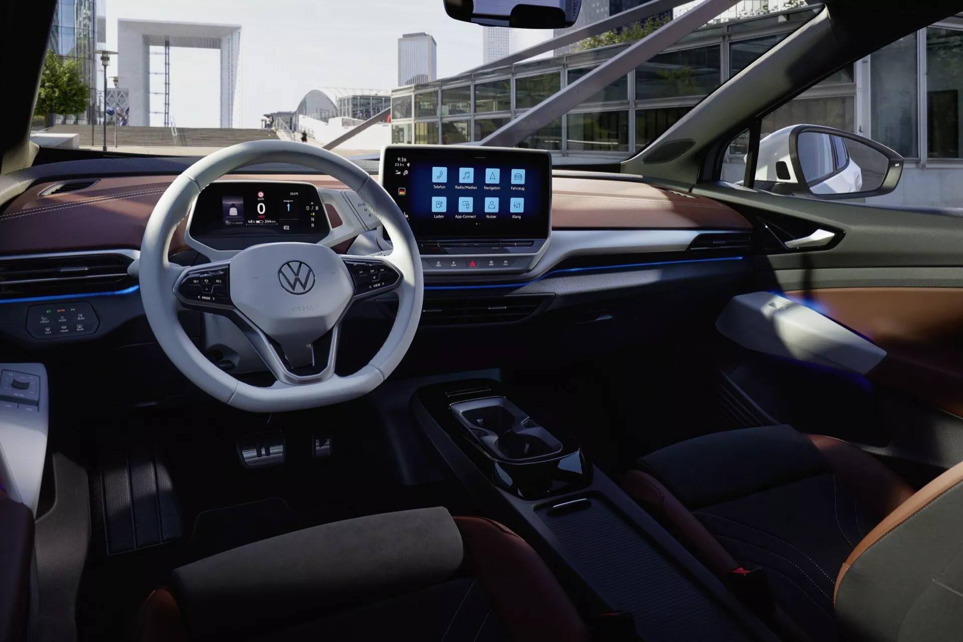 Interiérový záber modelu VW ID5 2022 (1. generácia) zo zadného sedadla, zvýrazňujúci luxus a technológie. Pohľad obsahuje dvojramenný volant s chrómovým lemovaním, digitálny prístrojový panel, veľký dotykový infotainment systém, modernú stredovú konzolu a mix kožených a drevených materiálov. Usporiadanie je navrhnuté s dôrazom na konektivitu s pokročilými funkciami, ako je Bluetooth a integrácia smartfónu, všetko osvetlené príjemným ambientným osvetlením pre útulnú atmosféru.