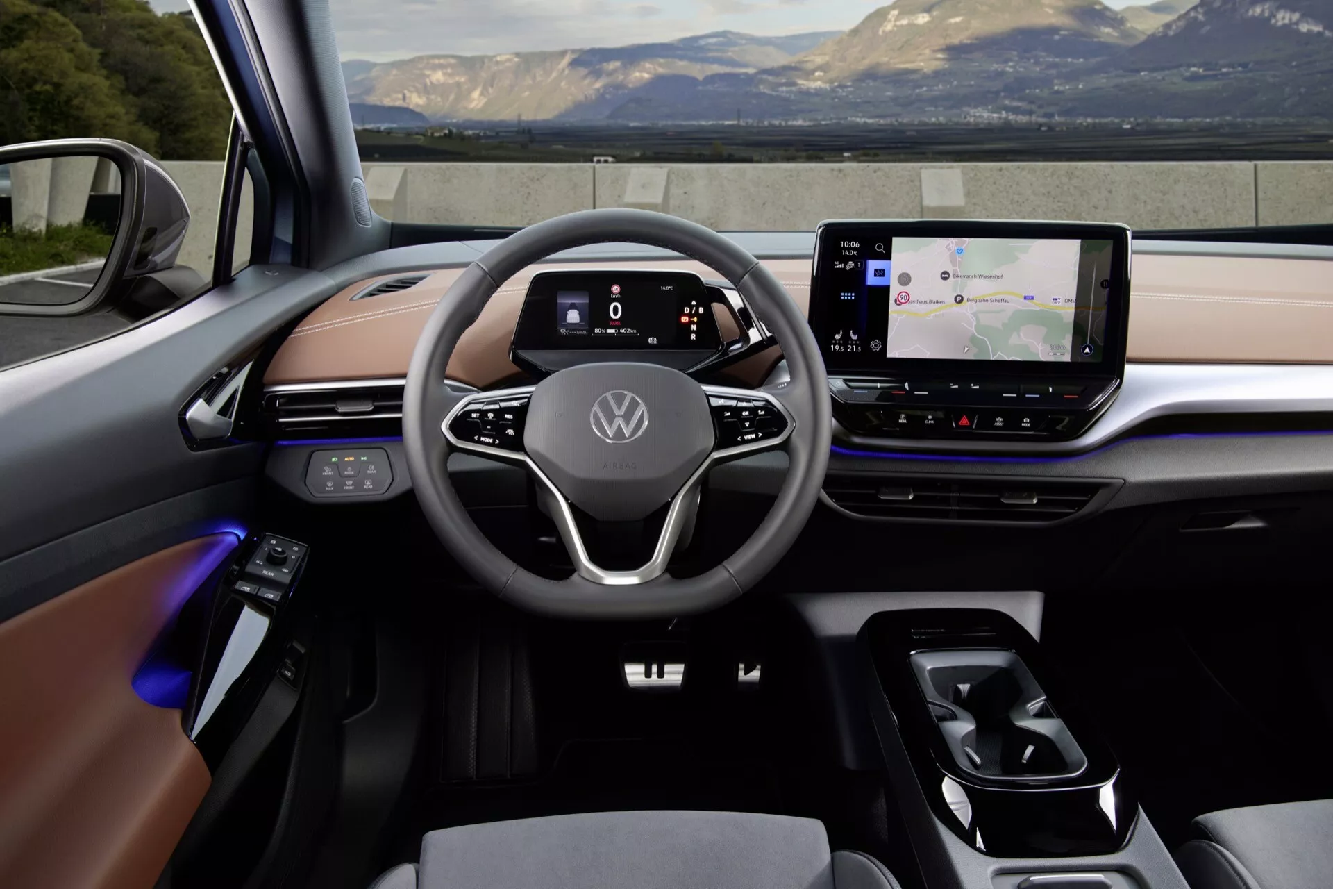 Pohľad na interiér moderného Volkswagen ID5 2022 (1. generácia) zo zadného sedadla. Palubná doska obsahuje veľký dotykový infotainment systém, prepínač prevodovky a plne digitálny prístrojový panel za minimalistickým dvojramenným volantom s dotykovými ovládacími prvkami. Interiér kombinuje kožu, mäkké plasty a kovové prvky s nastaviteľným ambientným osvetlením. Rozloženie naznačuje pokrokové prepojené vozidlo, pravdepodobne elektrické alebo hybridné.