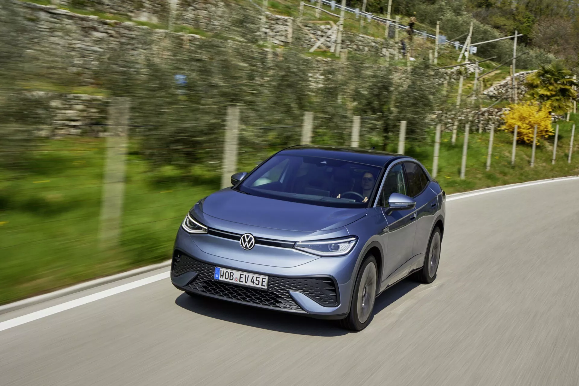 Obrázok zobrazuje moderný hatchback VW ID.5 2022 (1. generácia) jazdiaci po asfaltovej ceste. Zachytený z predného trojštvrťového pohľadu, zdôrazňuje prednú časť a stranu vodiča s jasným pohľadom na mriežku, svetlomety s LED denným svietením a hliníkové kolesá v pohybe. Karoséria má aerodynamické línie a elegantnú tmavú farbu umocnenú prirodzeným denným svetlom. Dynamický postoj auta je zdôraznený mierne nižším uhlom kamery a rozmazaným vidieckym pozadím, čo naznačuje rýchlosť.