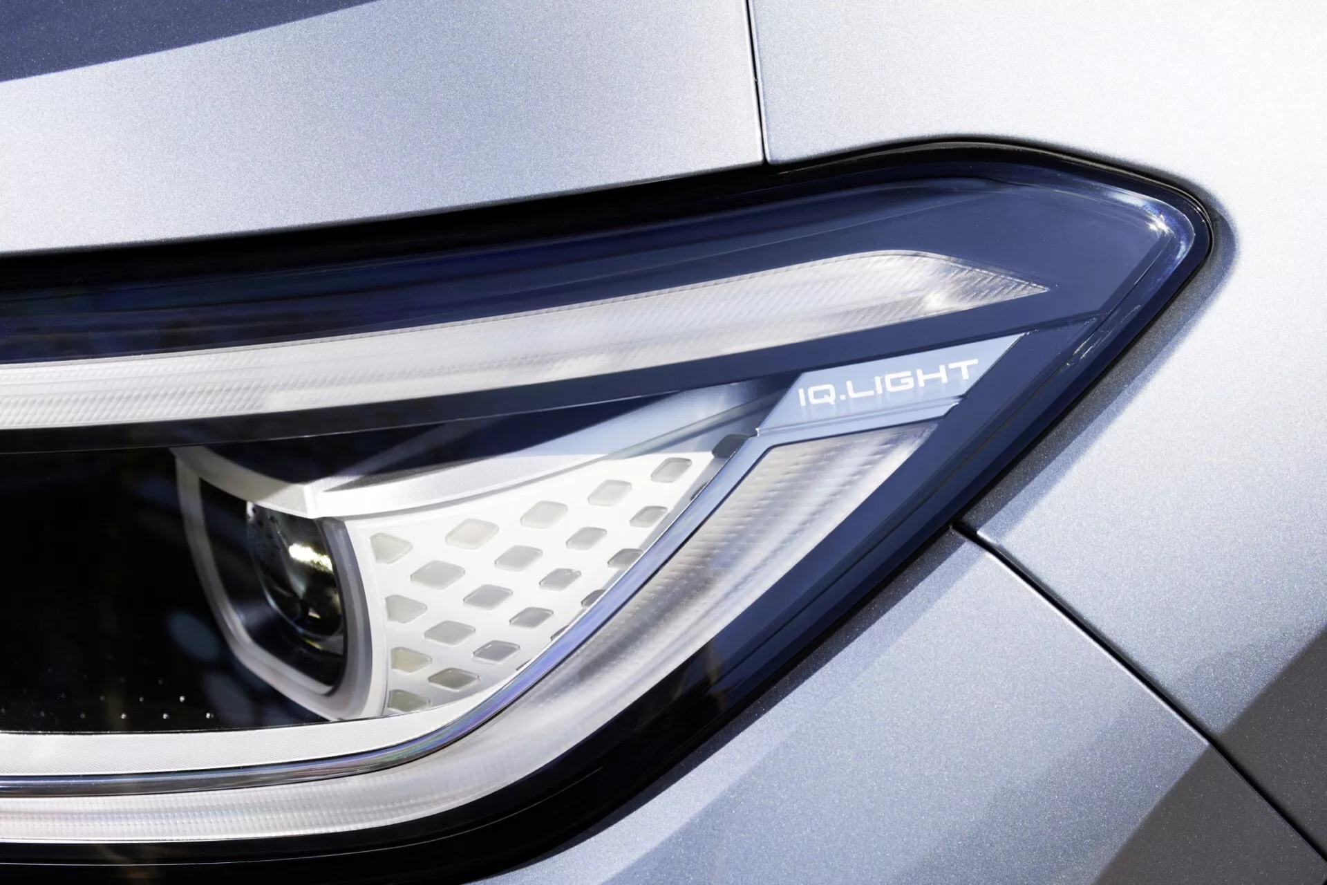 Detailný záber na svetlomet auta z bočného uhla, zobrazujúci zložitý inteligentný osvetľovací systém „IQ.LIGHT“, s elegantným a aerodynamickým dizajnom, geometrickým vzorom včelieho plástu a integráciou kovového panelu karosérie.