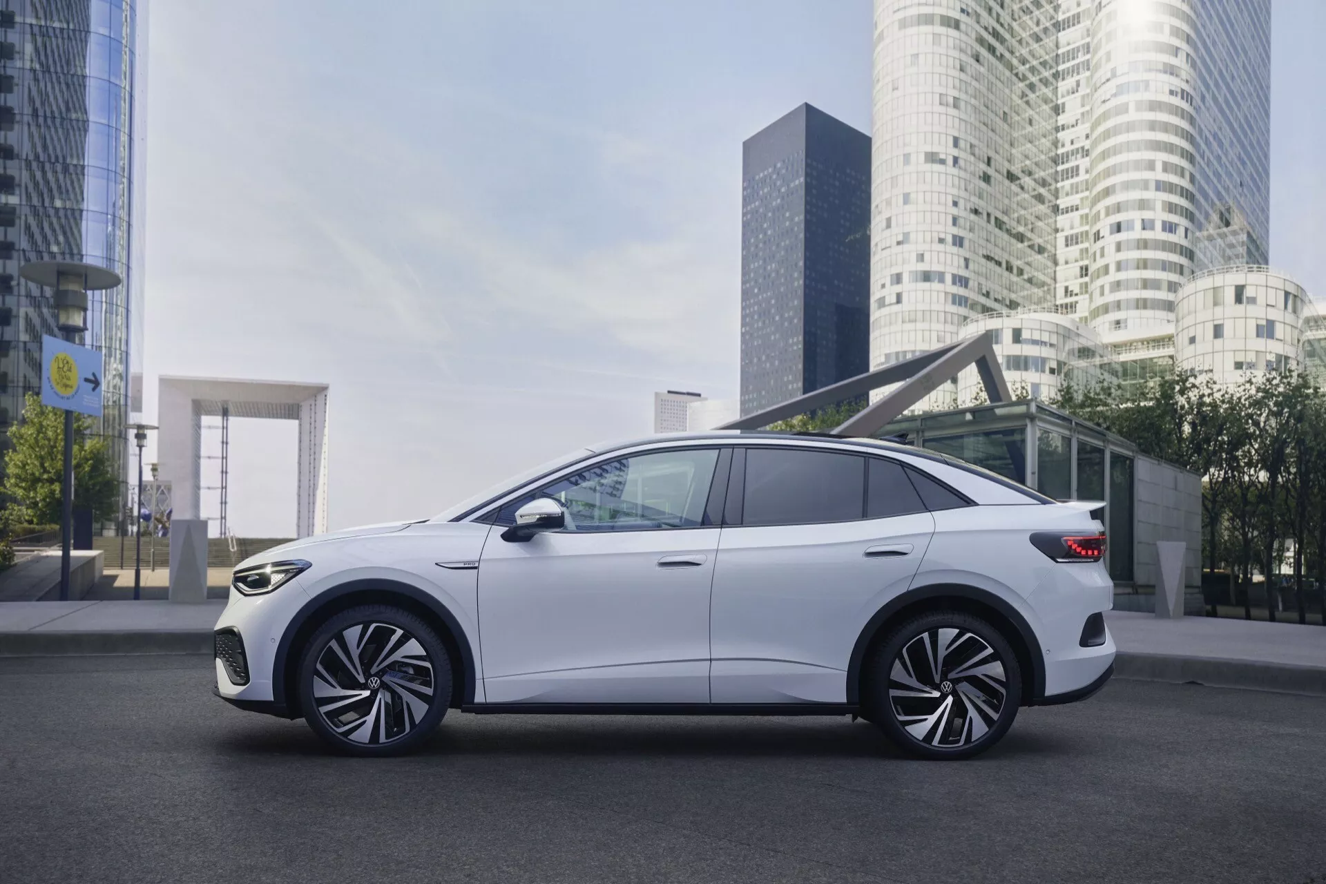 Obrázok VW ID.5 2022 prvej generácie zachytený v mestskom prostredí s vysokými budovami v pozadí. Bočný pohľad, mierne naklonený k prednému trojštvrťovému uhlu, zobrazuje veľké viacramenné zliatinové kolesá, elegantný hatchbackový dizajn, tenké LED svetlomety, integrované bočné zrkadlá s ukazovateľmi smeru, chrómové orámovanie okien, športový aerodynamický dizajn s výraznými líniami karosérie a metalické biele lakovanie.