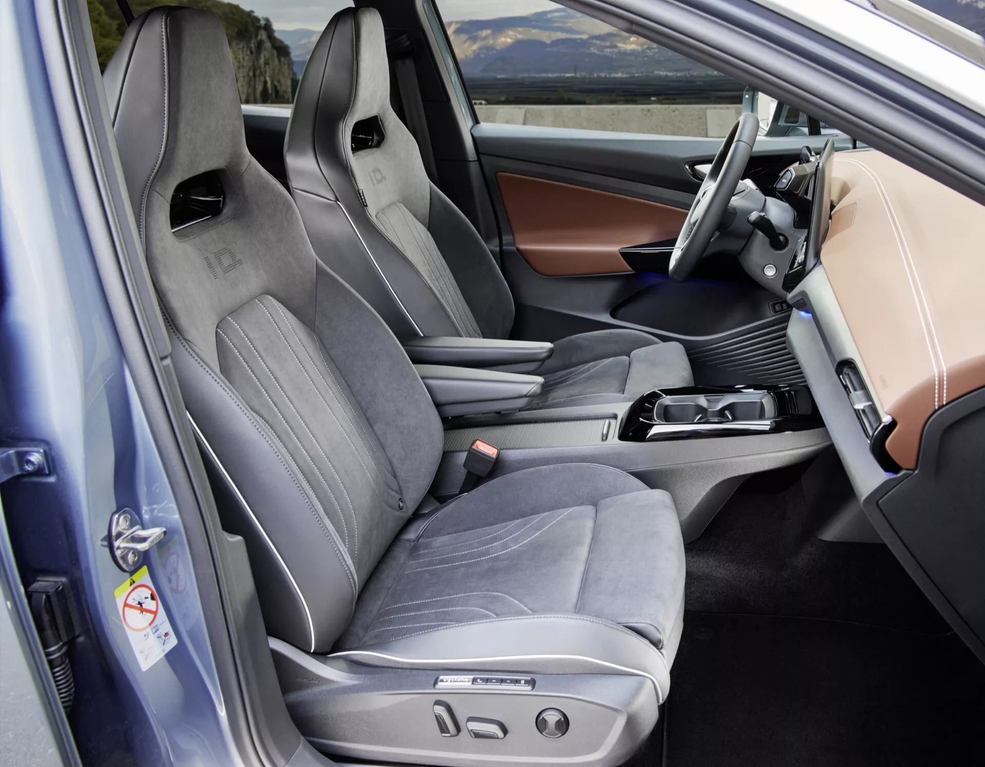 Pohľad do interiéru VW ID5 2022 (1. generácia) z pohľadu vodiča s otvorenými dverami, zachytávajúci prednú kabínu so zameraním na vodičovo sedadlo a časť sedadla spolujazdca. Zvýraznené prvky zahŕňajú športové vodičovo sedadlo s bočným podoprením, kombináciu kože a textilu a viacero elektrických ovládacích prvkov pre pohodlie. Elegantná centrálna konzola obsahuje radiacu páku pre automatickú prevodovku, ovládací gombík pre navigačný alebo multimediálny systém a malý úložný priestor, čo naznačuje použitie elektronickej parkovacej brzdy. Moderná prístrojová doska má displej pre infotainment a náhľad digitálneho prístrojového panela za volantom. Dverný panel obsahuje ovládacie prvky pre elektrické okna a zrkadlá s možnými nastaveniami pamäte pre polohy sedadiel. Celý interiér ukazuje pokročilé technológie a prémiové prvky, vhodné pre kategórie ako interiéry áut, automobilové technológie a interiéry luxusných vozidiel.