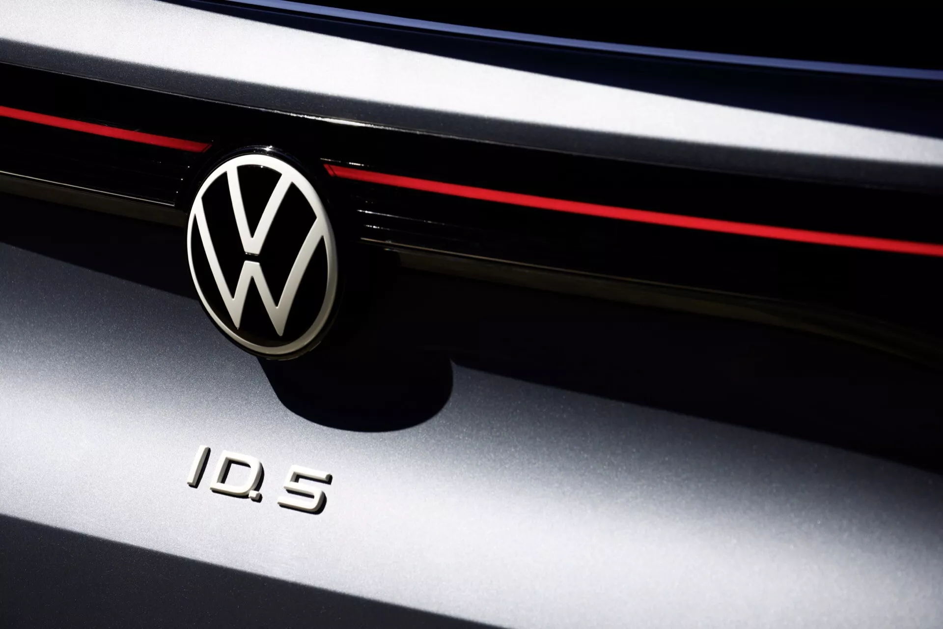 Záber na Volkswagen ID.5 2022 (1. generácia) s ikonickým chrómovým odznakom VW na čiernom kruhovom pozadí a označením modelu 'ID.5'. Obrázok zdôrazňuje prednú fasádu elektrického vozidla so štýlovou horizontálnou červenou čiarou, ktorá označuje športový dizajn. Záber s nízkou hĺbkou ostrosti zdôrazňuje detailné logá, pričom hra tieňov a svetla zvýrazňuje oblasť mriežky a okraj kapoty auta.
