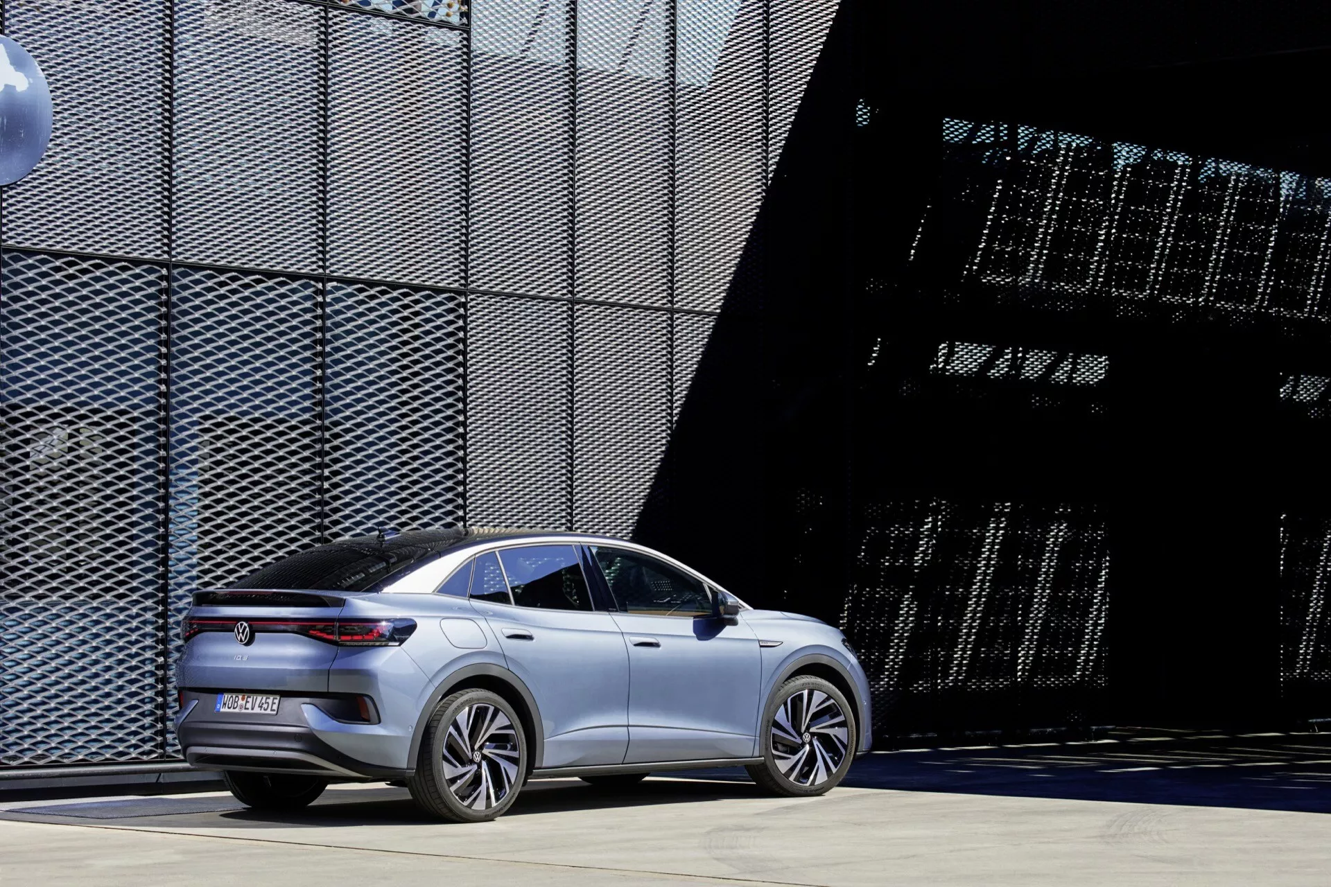Moderný, strieborný VW ID.5 2022 prvej generácie sedan so športovým, aerodynamickým dizajnom zaparkovaný vonku počas dňa, s architektonickými štruktúrami v pozadí. Uhol z troch štvrtín zozadu ukazuje elegantné línie karosérie, zapustené kľučky dverí, aerodynamické kolesá, zadné svetelné pruhy s LED technológiou, veľké hliníkové kolesá a sklonenú líniu strechy, čo naznačuje luxusný elektrický alebo hybridný model. Minimalistická estetika bez viditeľných výfukových rúrok, umiestnená na spevnenom povrchu s industriálnym, moderným mestským pozadím.