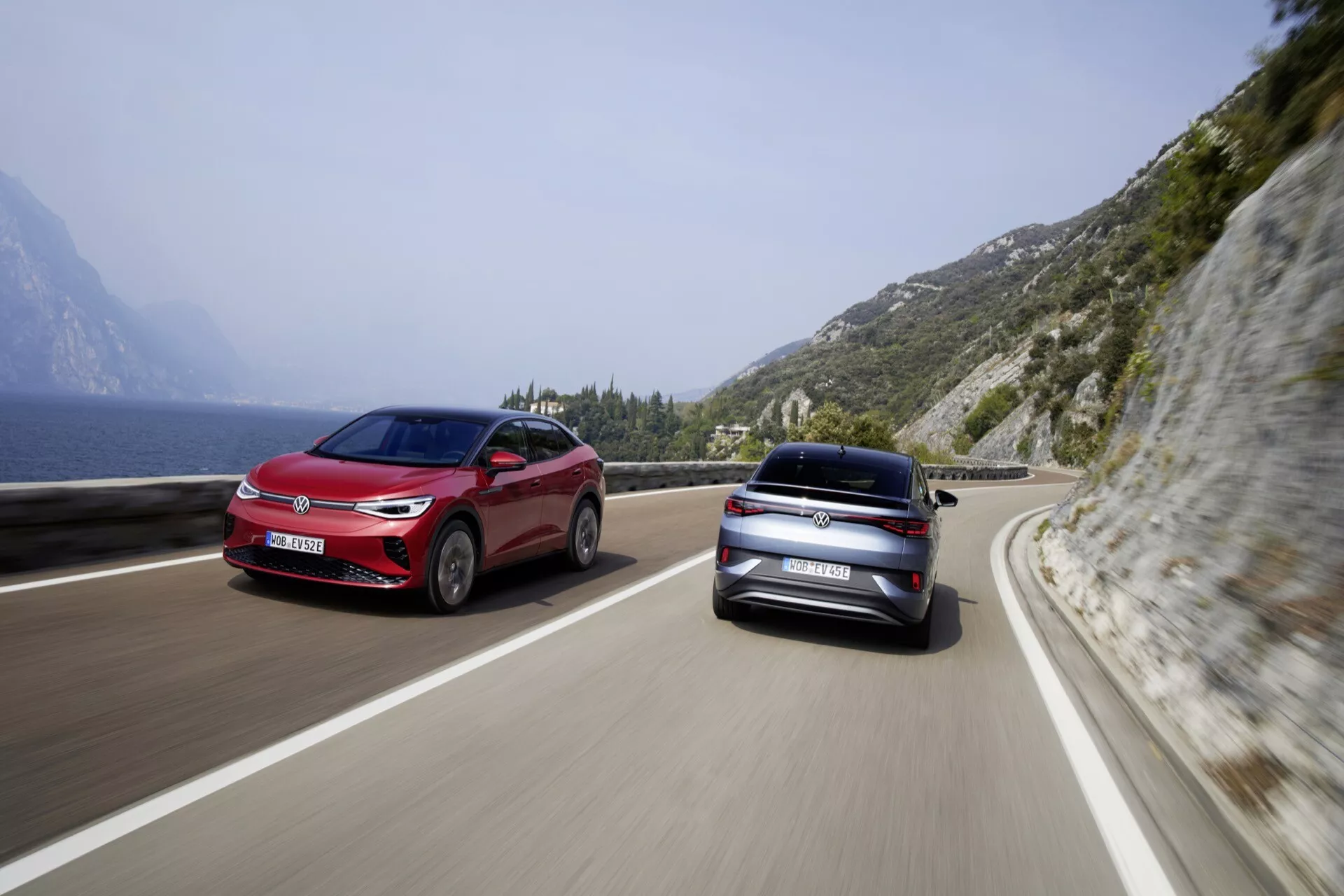 Obrázok ukazuje dve autá VW ID.5 2022 prvej generácie v pohybe na pobrežnej alebo horskej ceste pod jasnou oblohou a s kopcovitou, skalnatou krajinou vľavo. Oba vozidlá sú zachytené zo zadného trojštvrťového pohľadu, čo zdôrazňuje moderný, elegantný dizajn s viditeľnými zadnými svetlami, kufrom a zadným nárazníkom. Stredne veľké sedany alebo hatchbacky odrážajú lesklý povrch, čo naznačuje vysokú estetickú príťažlivosť. Scenéria evokuje pocit cestovania a slobody, čo naznačuje malebný a dynamický zážitok z jazdy.