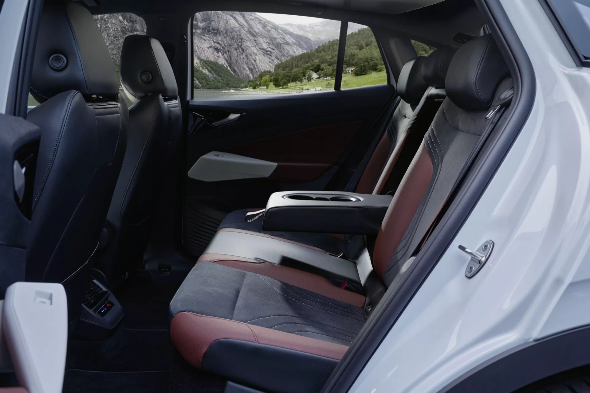 Pohľad do interiéru vozidla Volkswagen ID.5 z roku 2022 (prvá generácia), ktorý zachytáva zadnú pasažiersku časť z pravého uhla, zvýrazňujúc priestranné dvojfarebné sedadlá s tmavým a svetlým čalúnením, dobre integrovanú stredovú konzolu a otvorené pravé zadné pasažierske dvere zobrazujúce ovládacie prvky elektrických okien, zámkov dverí, mriežky reproduktorov a odkladací vrecko dverného panelu. Viditeľné sú koberce vozidla, pričom cez okná vidiek výhľad na malebné vonkajšie prostredie vyvoláva pocit pohody a dobrodružstva.