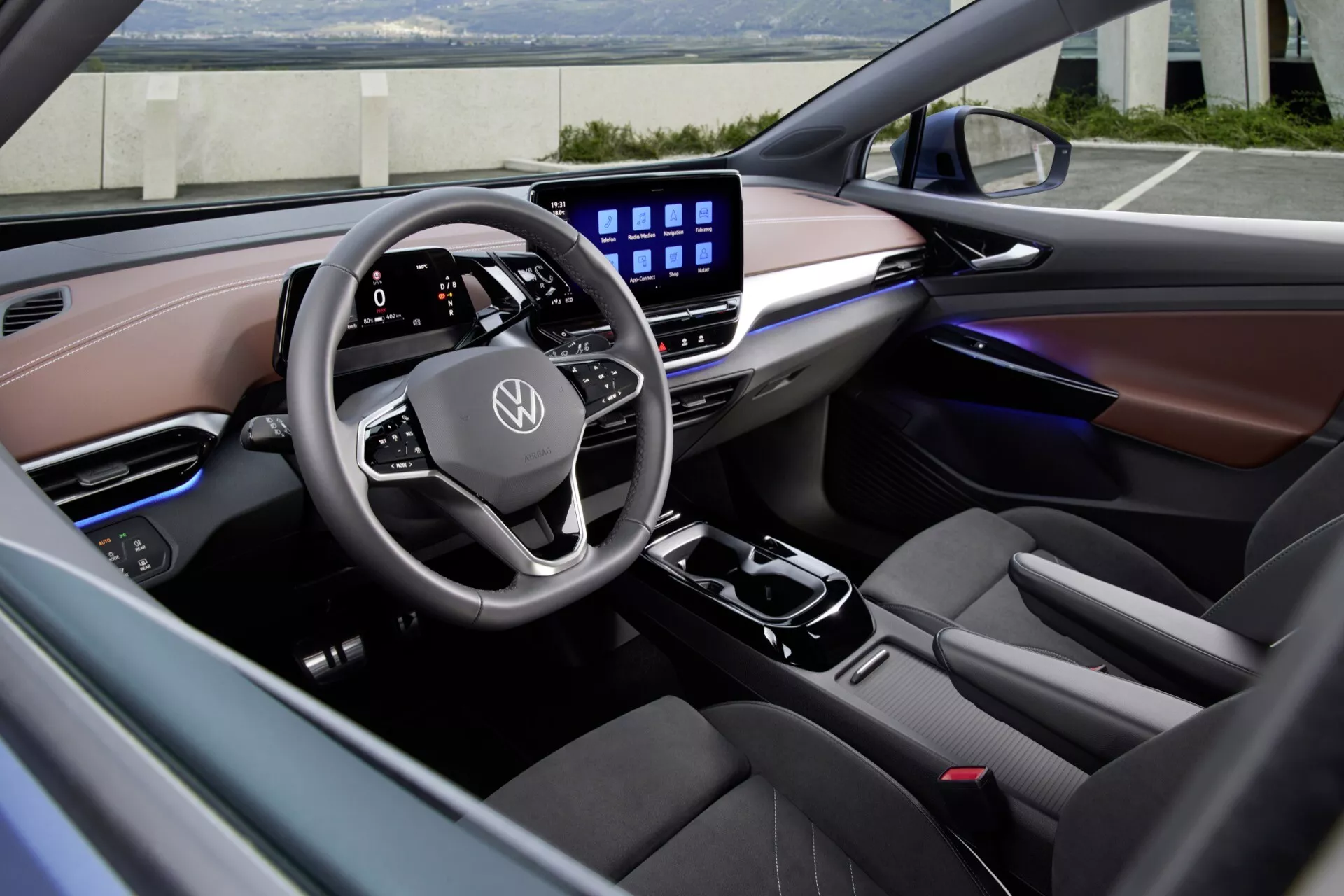 Interiér Volkswagen ID.5 2022 (1. generácia) zobrazený z ľavej strany, zameriavajúci sa na oblasť vodiča a palubnú dosku. Moderný multifunkčný volant s plochým spodkom a logom Volkswagen je obklopený digitálnym prístrojovým panelom a veľkým dotykovým displejom na stredovej konzole pre pokročilý infotainment systém. Palubná doska obsahuje kombináciu materiálov podobných koži v kontrastných svetlách a tmavých tónoch. Ambientné osvetlenie sa tiahne cez palubnú dosku a dverné panely, zdôrazňuje prémiovú kvalitu a luxusné dokončenie interiéru vozidla.