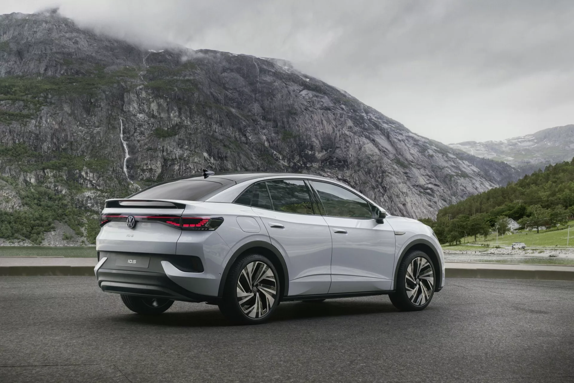 SUV VW ID5 2022 zachytený v modernom trojštvrtinovom pohľade zozadu. Obrázok zvýrazňuje pravú stranu vozidla z vyvýšeného uhla a ukazuje veľké kolesá z ľahkých zliatin s viacerými lúčmi, elegantné zadné LED svetlá, klesajúcu líniu strechy so zadným spojlerom, zatmavené okná, strieborný alebo svetlosivý lak s chrómovaným alebo leskle čiernym lemovaním. Vozidlo je umiestnené na suchom povrchu ciest s horami a vodopádom v pozadí, čo naznačuje dobrodružný a outdoorovo orientovaný vzhľad.