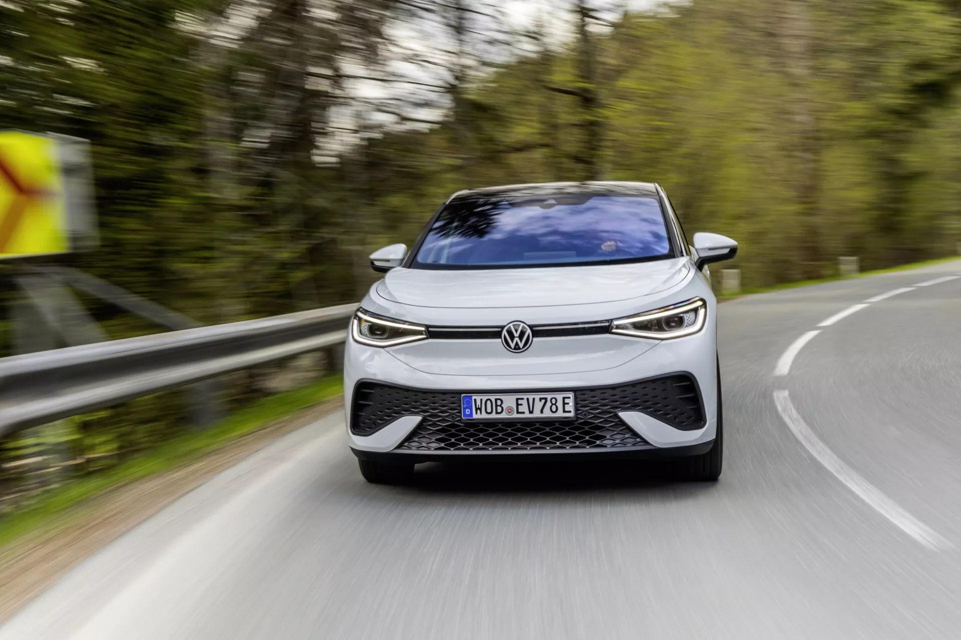 Volkswagen ID.5 2022 (1. generácia) zachytený na vidieckej ceste, ukazujúci nízky predný uhol so zameraním na elegantnú prednú časť vrátane mriežky, LED svetlometov a aerodynamického nárazníka. Auto sa pohybuje mierne doprava a dopredu, čo ukazuje jasné dizajnové a technologické prvky naznačujúce elektrické alebo hybridné vozidlo, registrované v Nemecku, s moderným dynamickým vzhľadom.