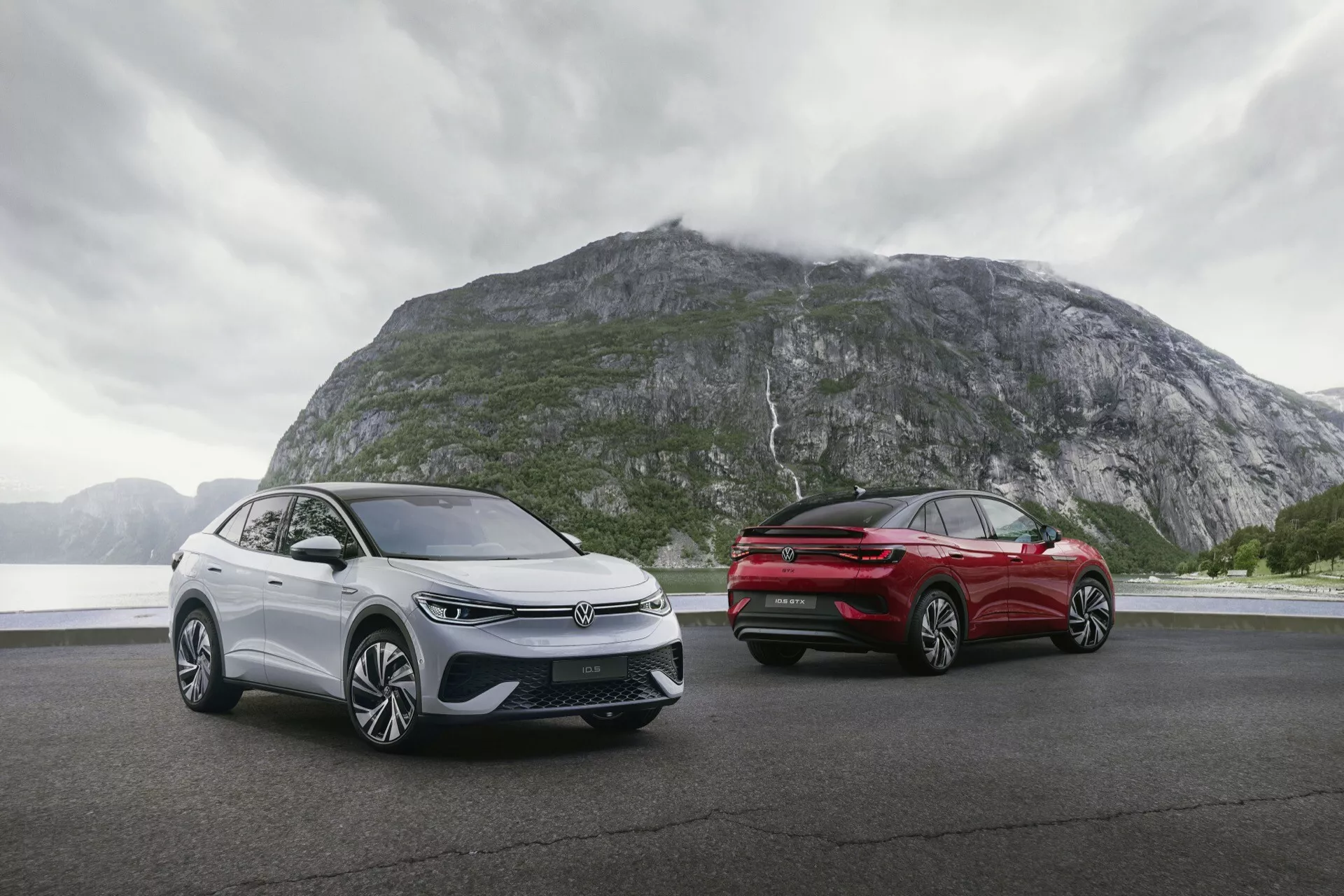 Dva vozidlá VW ID.5 2022 (1. generácia) zaparkované na rovnom povrchu s dramatickým horským pozadím. Ponuré počasie s dažďovými mrakmi pridáva na nálade. Vozidlo vľavo je zobrazené z trojštvrťového predného uhla s úzkymi LED svetlometmi, uzavretou mriežkou, senzormi ADAS a aerodynamickými kolesami. Vozidlo vpravo je viditeľné z trojštvrťového zadného uhla s úzkymi LED zadnými svetlami, zadným difúzorom a elegantným dizajnom. Obe vozidlá majú zliatinové kolesá, metalický povrch a elegantné bočné zrkadlá s integrovanými smerovkami.