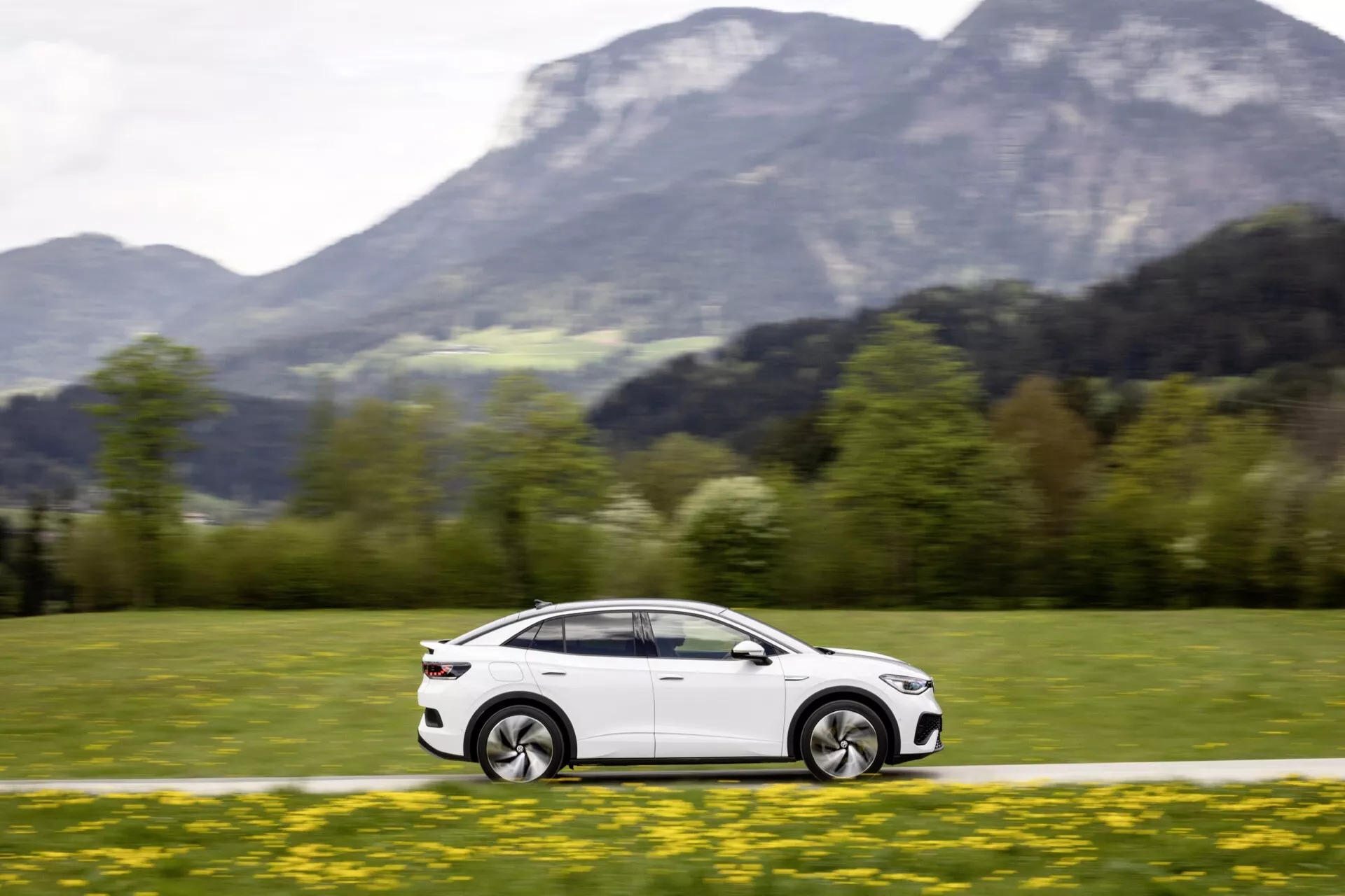 Obrázok zobrazuje dynamický bočný profil VW ID5 2022 (1. generácia) v pohybe, zdôraznený rozmazaním kolies a pozadia. Fotografia z nízkeho uhla a strednej vzdialenosti zvýrazňuje aerodynamický a elegantný dizajn auta, vrátane dverí, bočných okien, zrkadiel a dizajnových línií pozdĺž strany. Kolesá sú v pohybe a mierne natočené, čo naznačuje manéver alebo zákrutu. Odrážanie svetla ukazuje na lesklý, dobre udržiavaný exteriér. Auto je zasadené do malebnej vonkajšej krajiny so zelenou trávou v popredí a horami v pozadí, čím sa zvyšuje pocit rýchlosti a dobrodružstva.