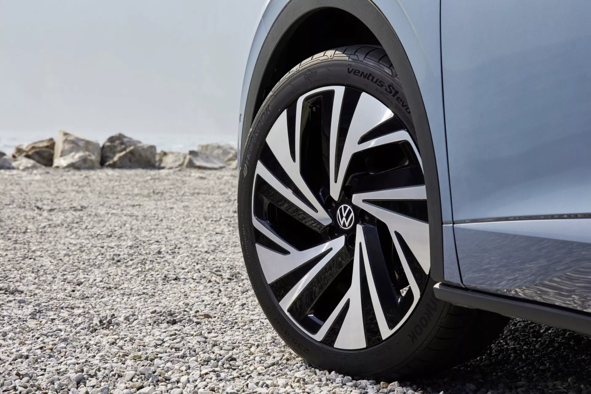 Záber z nízkeho uhla zblízka na predné koleso na strane spolujazdca automobilu Volkswagen ID.5 2022 (1. generácia). Koleso s viacerými lúčmi má dvojfarebnú schému s čiernymi a striebornými alebo bielymi akcentmi, ktoré zdôrazňujú jeho moderný, dynamický vzhľad. Logo Volkswagen je výrazne zobrazené na strede krytu. Pneumatika nesie nápis 'Continental', čo naznačuje vysoko kvalitnú značku. Obrázok, zachytený vonku na oblázkovom povrchu s veľkými kameňmi v pozadí, zdôrazňuje koleso s ostrým zameraním, ktoré sa postupne rozmazáva smerom k telu vozidla a pozadiu, naznačujúcim pobrežné alebo horské prostredie za denného svetla.