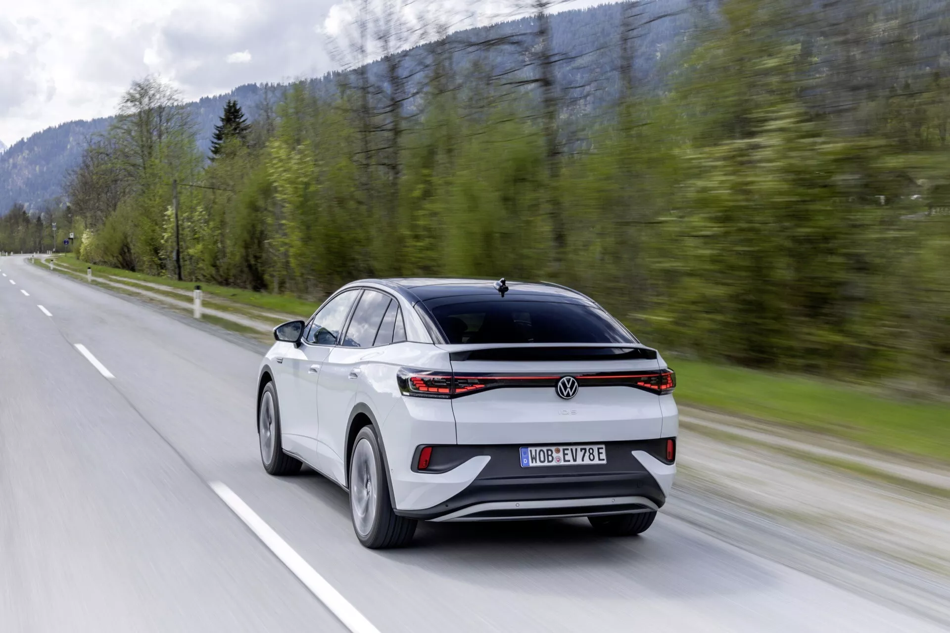 2022 VW ID.5 prvej generácie moderný crossover/SUV, zachytený z troch štvrtín zadného pohľadu, ktorý zobrazuje športový a aerodynamický dizajn so sklopeným zadným oknom, charakteristickou LED zadnou svetelnou lištou, dvojfarebným kontrastným vzhľadom v bielej a čiernej farbe, prémiovými viacramennými zliatinovými kolesami, snímačmi pokročilého asistenčného systému vodiča, centrálnou registračnou značkou a logom, zachytený v pohybe na dvojprúdovej vidieckej ceste s lesnatým horským pozadím, čo evokuje rýchlosť a výkon, pravdepodobne elektrické alebo hybridné vozidlo.