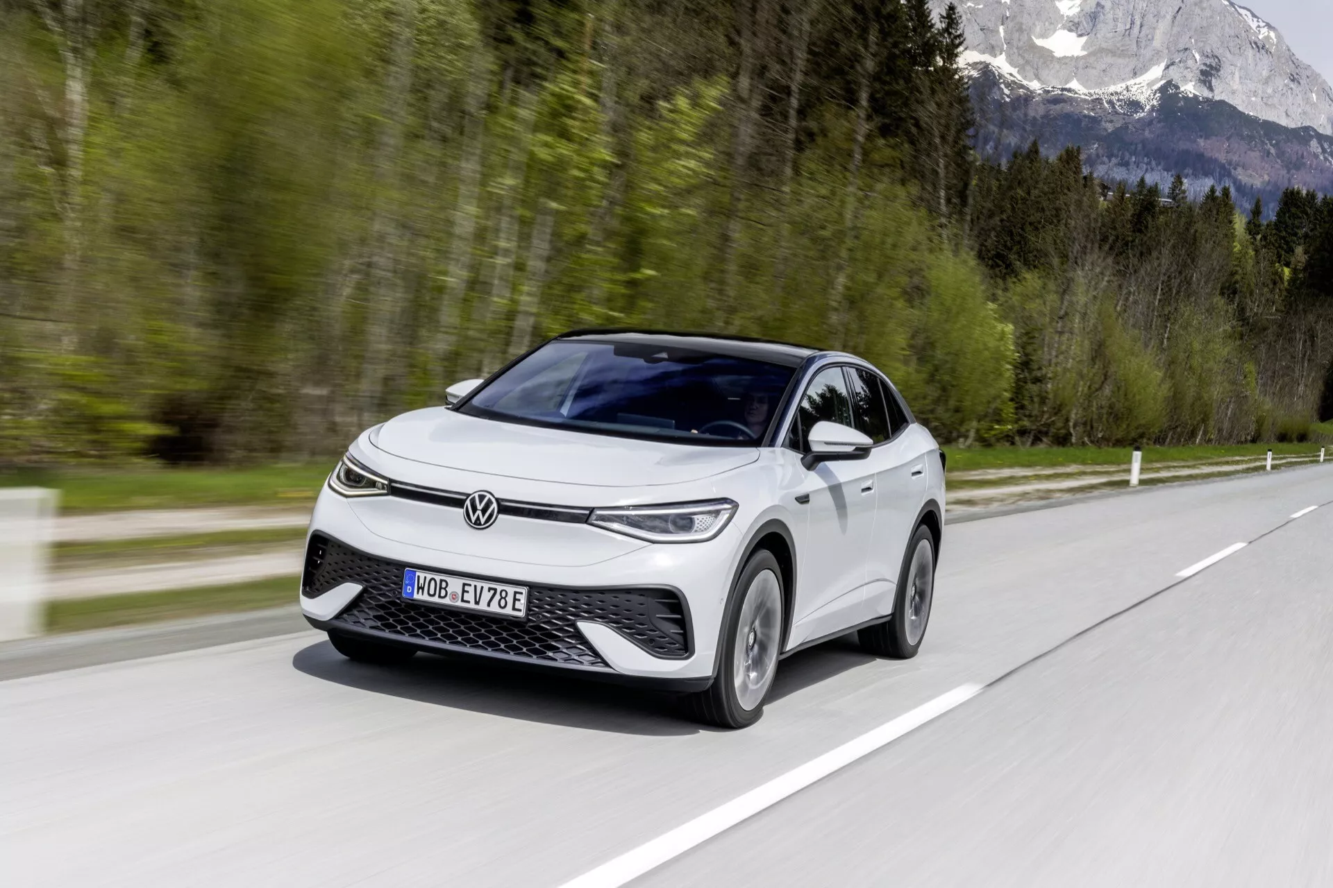Obraz moderného VW ID.5 2022 (1. generácia) elektrického vozidla, zachyteného za denného svetla počas jazdy, zobrazujúci pohľad z predného a bočného profilu pod uhlom 45 stupňov. Auto, hatchback alebo kompaktné SUV, je zdôraznené svojou výraznou prednou mriežkou a úzkymi horizontálnymi svetlometmi. Vozidlo vykazuje vlastnosti ako denné svietenie, aerodynamické ráfiky, bočné zrkadlá s integrovanými smerovými svetlami, panoramatickú strechu a dvojfarebné lakovanie. Rozmazané pozadie a povrch cesty na fotografii zvýrazňujú pohyb vozidla, zatiaľ čo 'E' na evidenčnom čísle ho identifikuje ako elektrický model.
