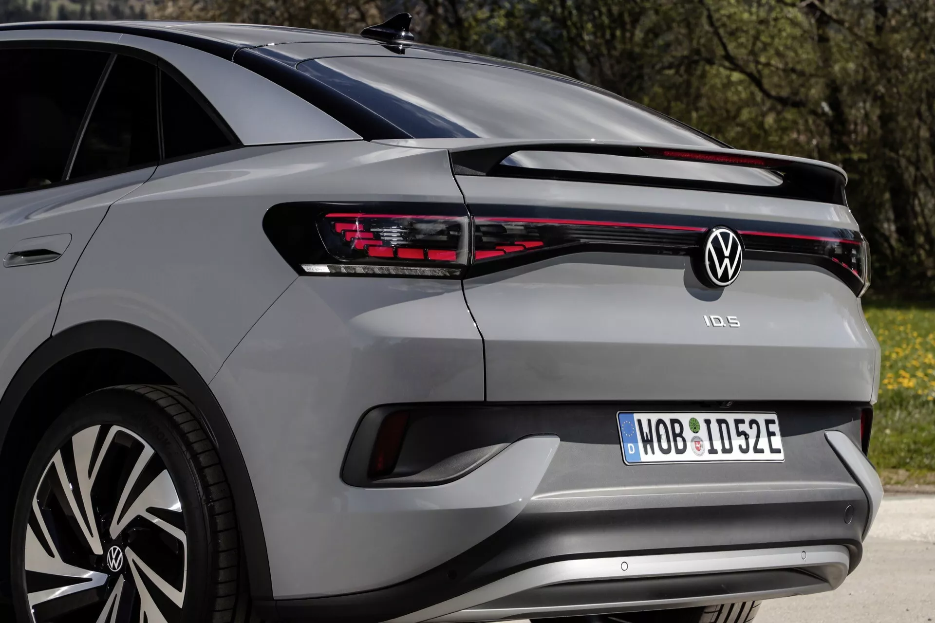 Zadný trojštvrťový uhol pohľadu na moderný hatchback VW ID.5 2022 (1. generácia) ukazujúci LED zadné svetlo, ktoré sa tiahne po celej šírke, výrazné segmentované zadné svetlá na oboch stranách, centrálny znak značky, názov modelu pod svetlom, registračnú značku v jej výklenku, zadný nárazník so senzormi parkovania, štýlový zadný spojler s namontovaným stieračom a časť viaclúčového hliníkového kolesa, odrážajúci exteriérové dizajnové prvky a technológie.