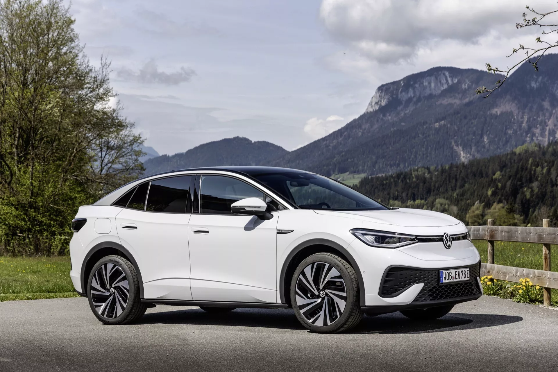 Moderné biele SUV VW ID.5 z roku 2022 zaparkované vonku v malebnom prostredí so zeleňou a horami. Vozidlo je zachytené z trojštvrťového predného pohľadu, zvýrazňujúc prednú mriežku, LED svetlá, zliatinové kolesá a šikmú líniu strechy. Obraz naznačuje pokročilé technologické funkcie, ako sú kamerové bočné zrkadlá, a prémiový, aerodynamický dizajn. Malebné prostredie podporuje vhodnosť auta pre mestské aj vidiecke prostredie.