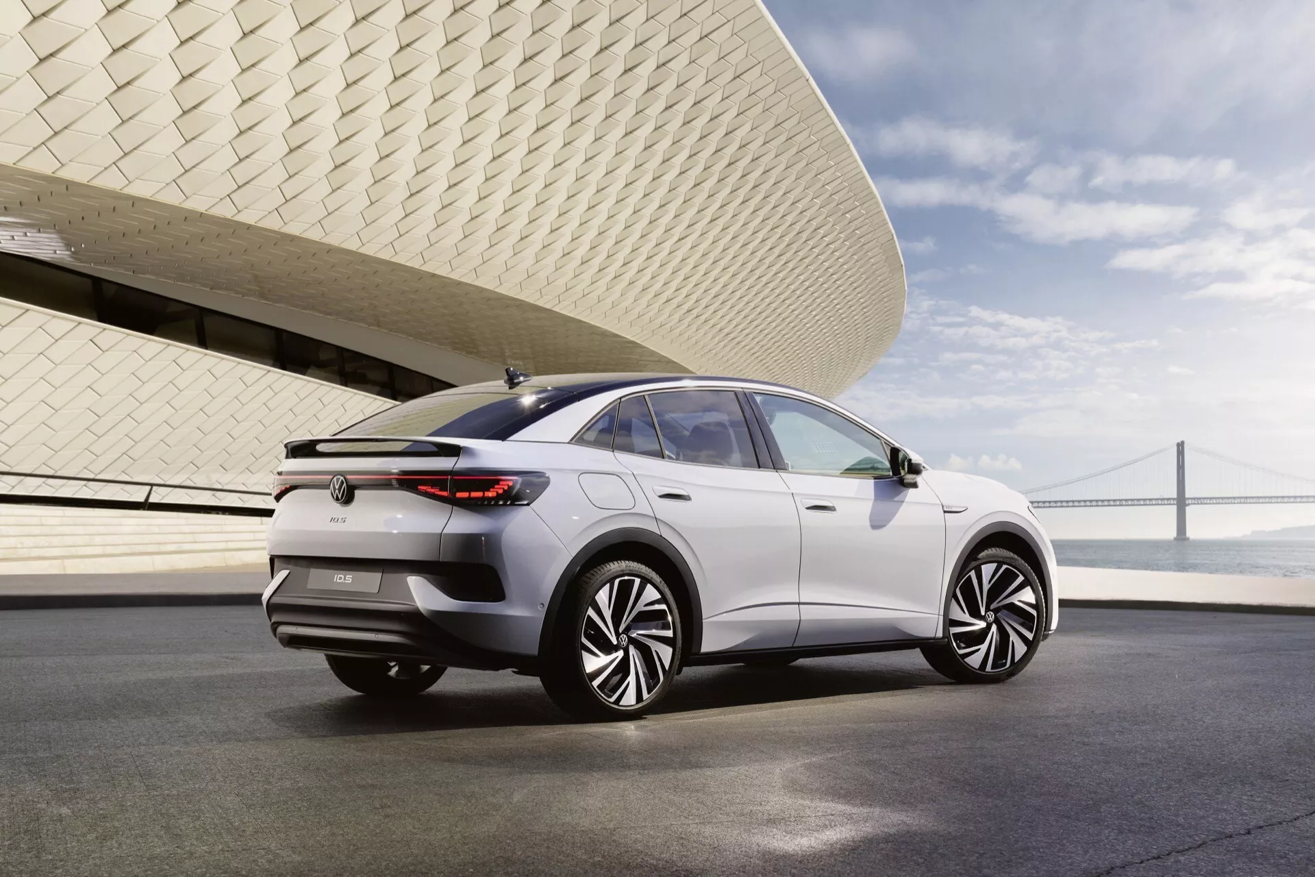 Zadný trojštvrťový pohľad na VW ID.5 2022 (1. generácia) ukazuje jeho elegantný a moderný dizajn. Crossover/SUV s klesajúcou líniou strechy, veľkými kolesami s prepracovaným viacramenným dizajnom, zapustenými kľučkami dverí, úzkymi horizontálnymi zadnými svetlami, minimalistickým zadným nárazníkom, panoramatickou strechou a spätnými zrkadlami so zabudovanými smerovými svetlami. Auto je vo svetlej farbe, pravdepodobne strieborné alebo biele. Fotografia je vyhotovená v mestskom prostredí s mostom a modernou budovou v pozadí, za jasného prirodzeného osvetlenia.