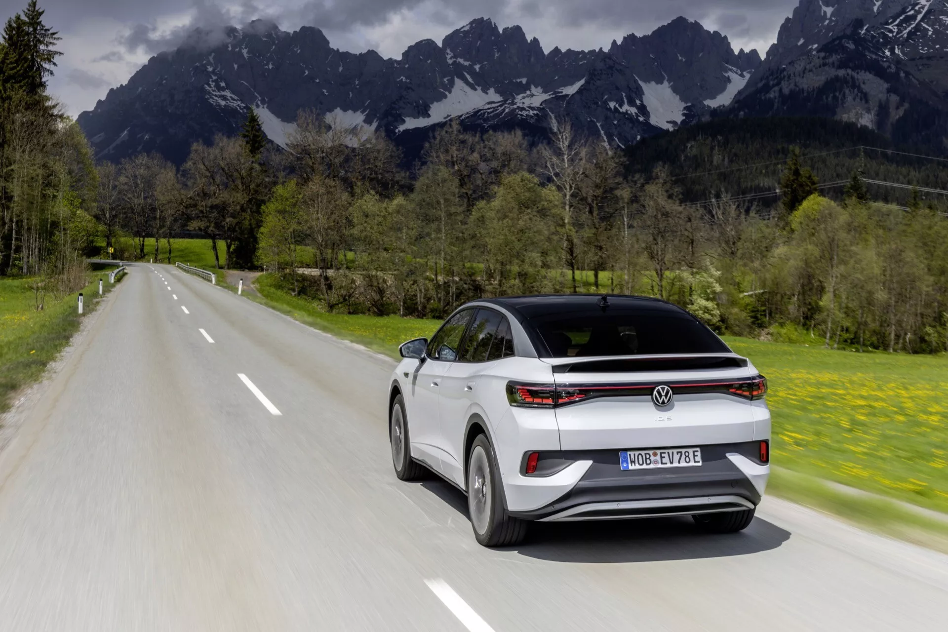 Elektrické SUV Volkswagen ID.4 z roku 2022 zachytené v pohybe z uhla zadnej tretiny, zobrazujúce jeho moderný aerodynamický dizajn na pozadí prírodnej krajiny. Vozidlo, umiestnené v strede rámu, ide po vidieckej dvojpruhovej ceste obklopenej bujnou zeleňou a vzdialenými horami. Výrazné črty zahŕňajú jeho šikmú strechu, horizontálne zadné svetlá a nápadné logo Volkswagen. Obrázok zdôrazňuje dynamický dizajn a elektrickú povahu SUV za prirodzených denných podmienok s ľahkým rozostrením pohybu na zdôraznenie pohybu.