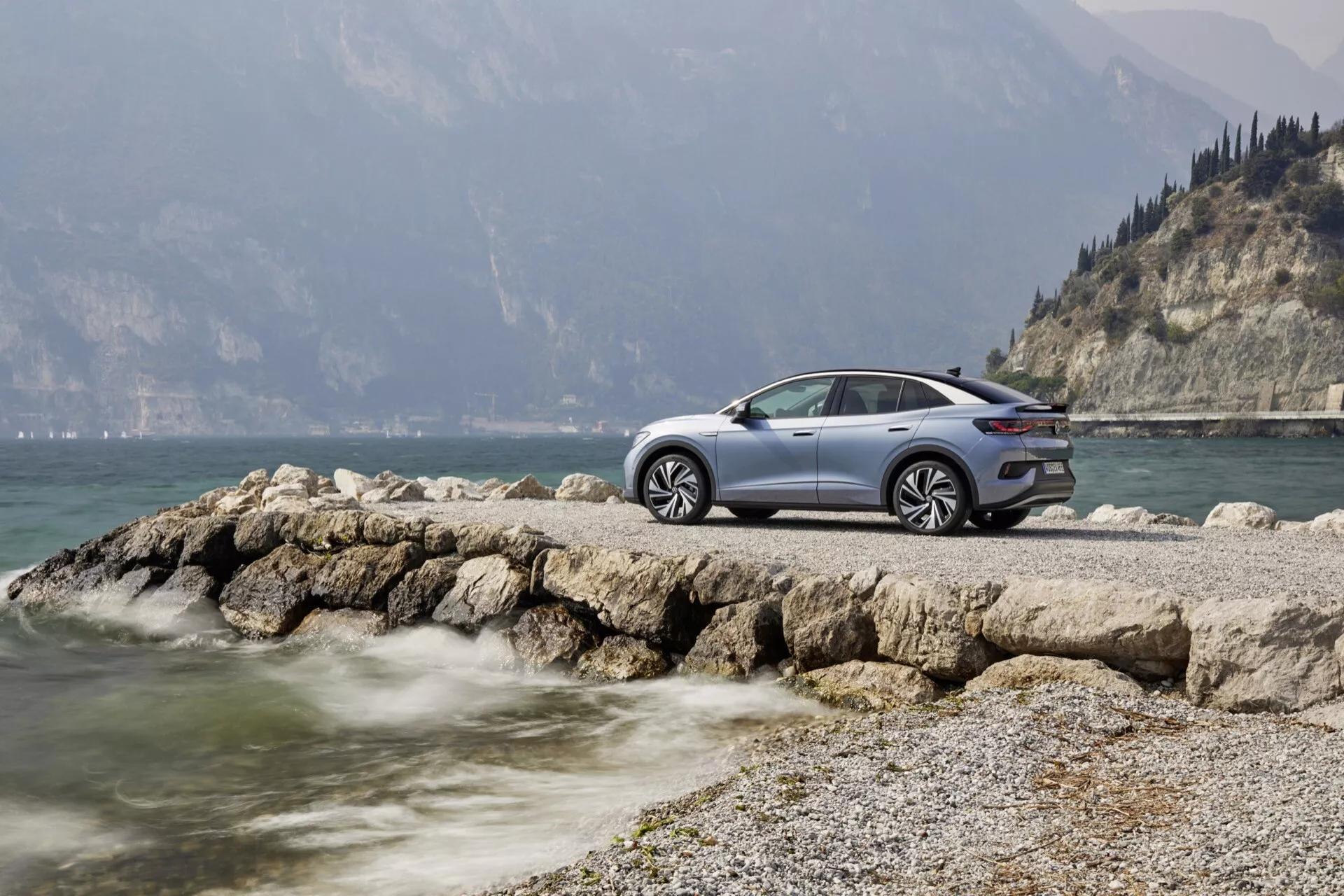 Auto VW ID5 2022 (1. generácia) zaparkované na skalnatom výbežku pri vodnej ploche s horským pozadím, čiastočne zakrytým hmlou. Bočný pohľad na stranu vodiča s vysokou svetlou výškou, veľkými diskovými kolesami, LED svetlami, elegantnými líniami karosérie a metalickým lakom. Obrázok vyvoláva pocit luxusu, moderného dizajnu a dobrodružstva v pokojnom prírodnom prostredí.