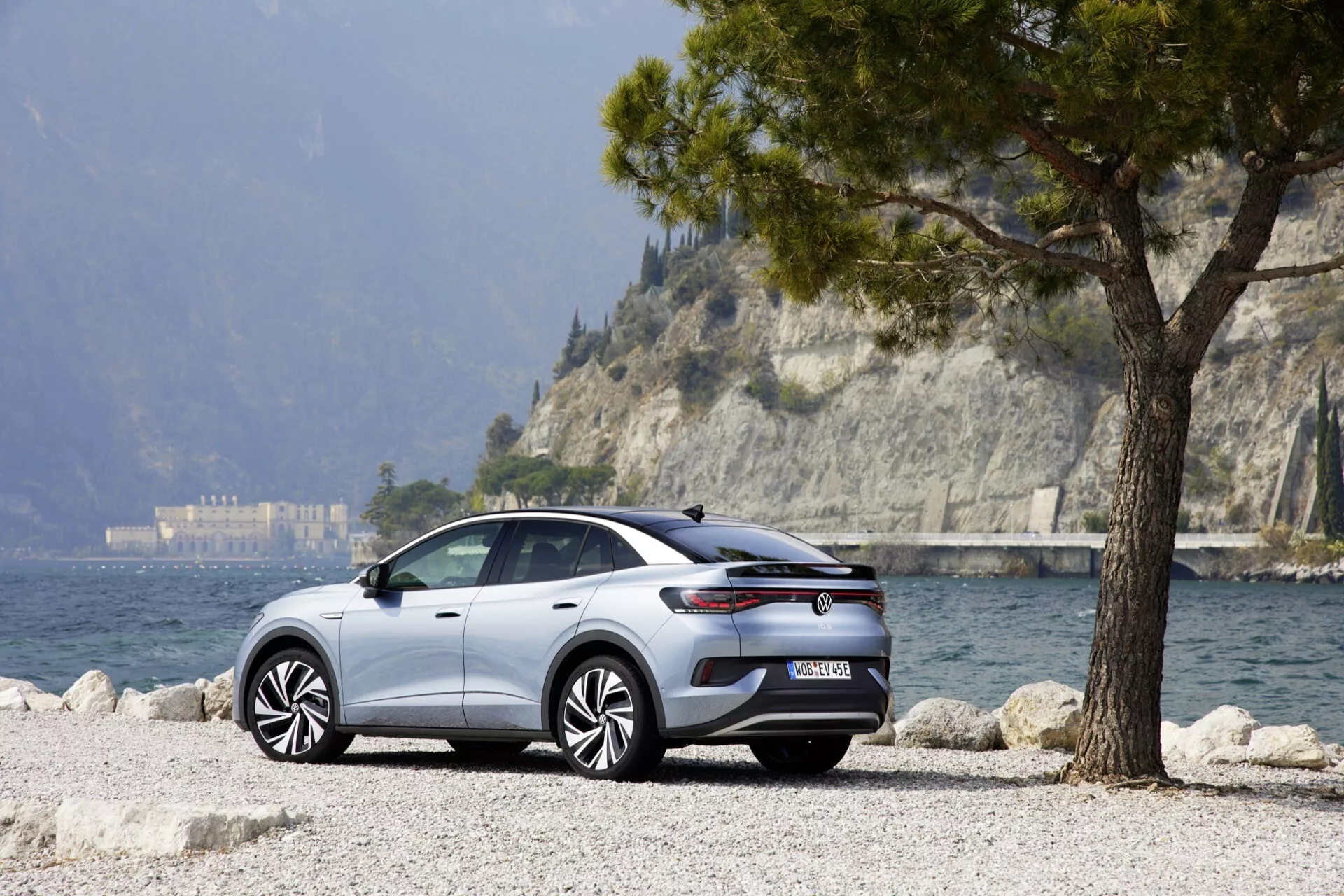 Moderný VW ID.5 2022, prvá generácia, zaparkovaný na malebnom vonkajšom mieste pri jazere. Pohľad zo zadnej troch štvrtín zvýrazňuje zadný a bočný profil vozidla, s viacramennými zliatinovými kolesami, zadnými svetlami, bočnými zrkadlami, kľučkami dverí a elegantnými líniami. Štrkový povrch a strom naznačujú jeho úžitkovú alebo crossover/SUV povahu. Dobre osvetlený za denného svetla, obraz naznačuje pokročilé technológie vozidla ako asistenčné systémy pre vodičov a funkcie konektivity.