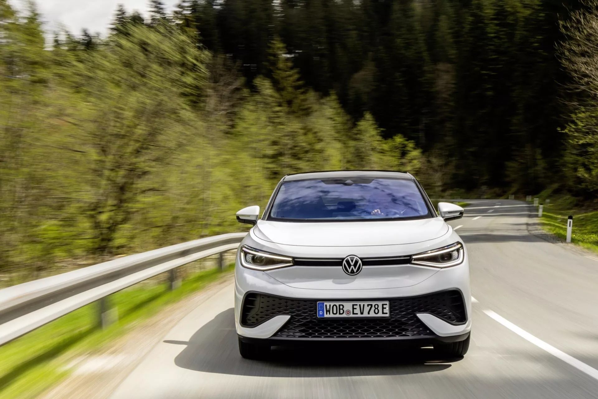 Obrázok zobrazuje Volkswagen ID.5 2022 (1. generácia) z predného, mierne posunutého uhla, ako sa blíži k fotoaparátu. Moderný dizajn auta zahŕňa nízky, široký postoj s výrazne zakrivenou kapotou, tenkou horizontálnou mriežkou, LED svetlami a veľkými nasávacími otvormi v prednom nárazníku, čo naznačuje výkonnostne orientovaný alebo agresívny dizajn. Viditeľná ŠPZ naznačuje, že automobil je na verejnej komunikácii a rozmazanie pohybu okolo vozidla a pozadia naznačuje, že auto je v pohybe v prírodnom, snáď lesnatom prostredí počas dňa. Písmeno 'E' na ŠPZ naznačuje, že ide o elektrické alebo hybridné vozidlo.
