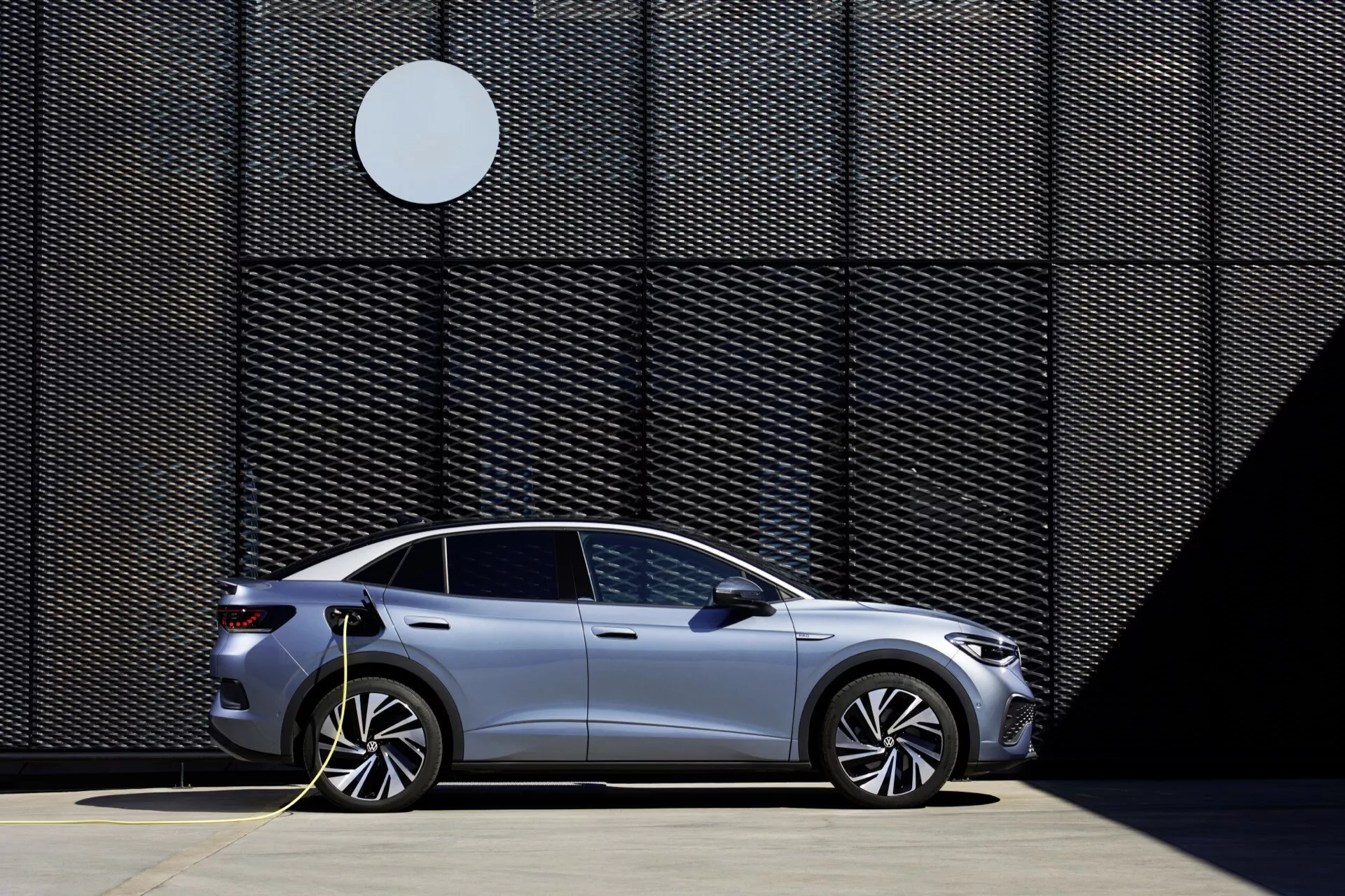 Bočný profil strieborného elektrického vozidla VW ID.5 2022 zaparkovaného a pripojeného k nabíjacej stanici. Vozidlo má aerodynamické kolesá, štyri dvere a moderný, elegantný dizajn, všetko zdôraznené jasným, slnečným dňom. Na pozadí obrázku je geometrická stena s kontrastným kruhovým tvarom, čo zdôrazňuje umiestnenie vozidla v určenej oblasti pre nabíjanie elektromobilov.