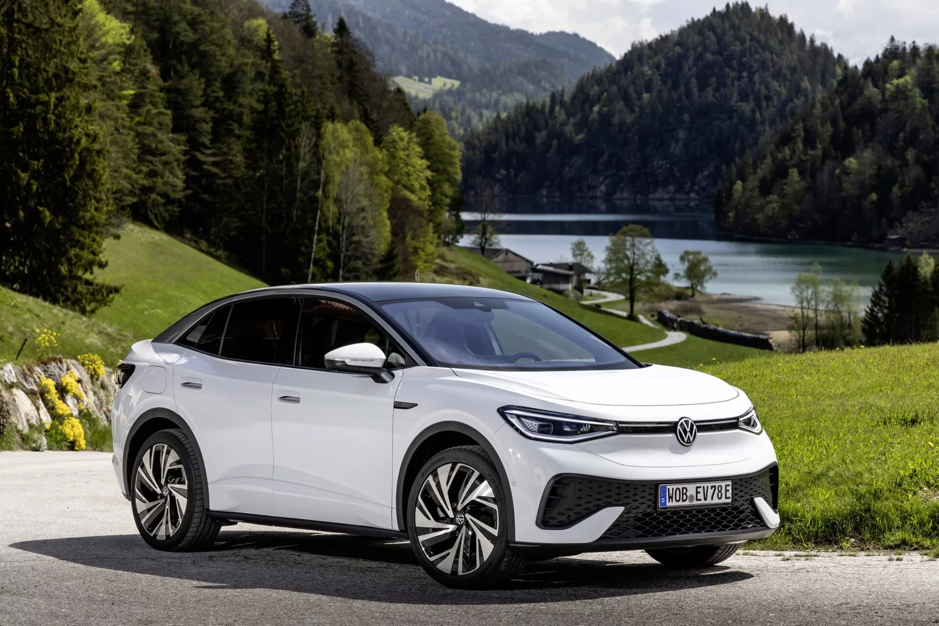 Obrázok bieleho modelu VW ID.5 z roku 2022 zaparkovaného na asfaltovej ceste s pokojnou prírodou v pozadí, vrátane zelenej trávy, stromov a jazera. Snímka z nízkeho uhla zo štvrťového predného pohľadu zvýrazňuje úzke LED svetlomety, výrazný stredový emblém, uzavretú masku, integrovaný predný nárazník s prívodom vzduchu a pravdepodobne parkovacími senzormi alebo adaptívnym tempomatom. Bočný profil ukazuje aerodynamicky navrhnuté viacpaprskové zliatinové disky, klesajúcu líniu strechy kombinujúcu štýly SUV a kupé, zrkadlá so zabudovanými smerovkami, zapustené kľučky dverí naznačujúce bezkľúčový vstup, chrómové alebo kovové okenné lišty a panoramatickú strechu. Absencia výfukových potrubí naznačuje, že vozidlo je elektrické. Prirodzené, dobre vyvážené denné svetlo a ostré zaostrenie zvýrazňujú ekologický dizajn vozidla a jeho estetickú príťažlivosť vonkajšieho prostredia.