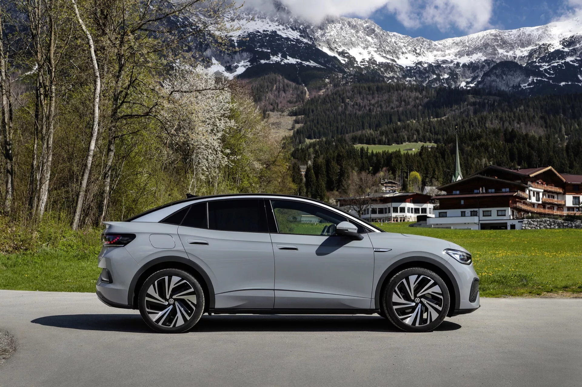 Bočný profil strieborno-šedého VW ID.5 z roku 2022 (1. generácia), predstavujúci elegantný a aerodynamický dizajn s výraznými obrysovými líniami, modernými zliatinovými kolesami, farebnými bočnými zrkadlami a zarovnanými kľučkami dverí, zaparkovaný na dláždenej ploche s malebným horským pozadím a domom v štýle chaty.