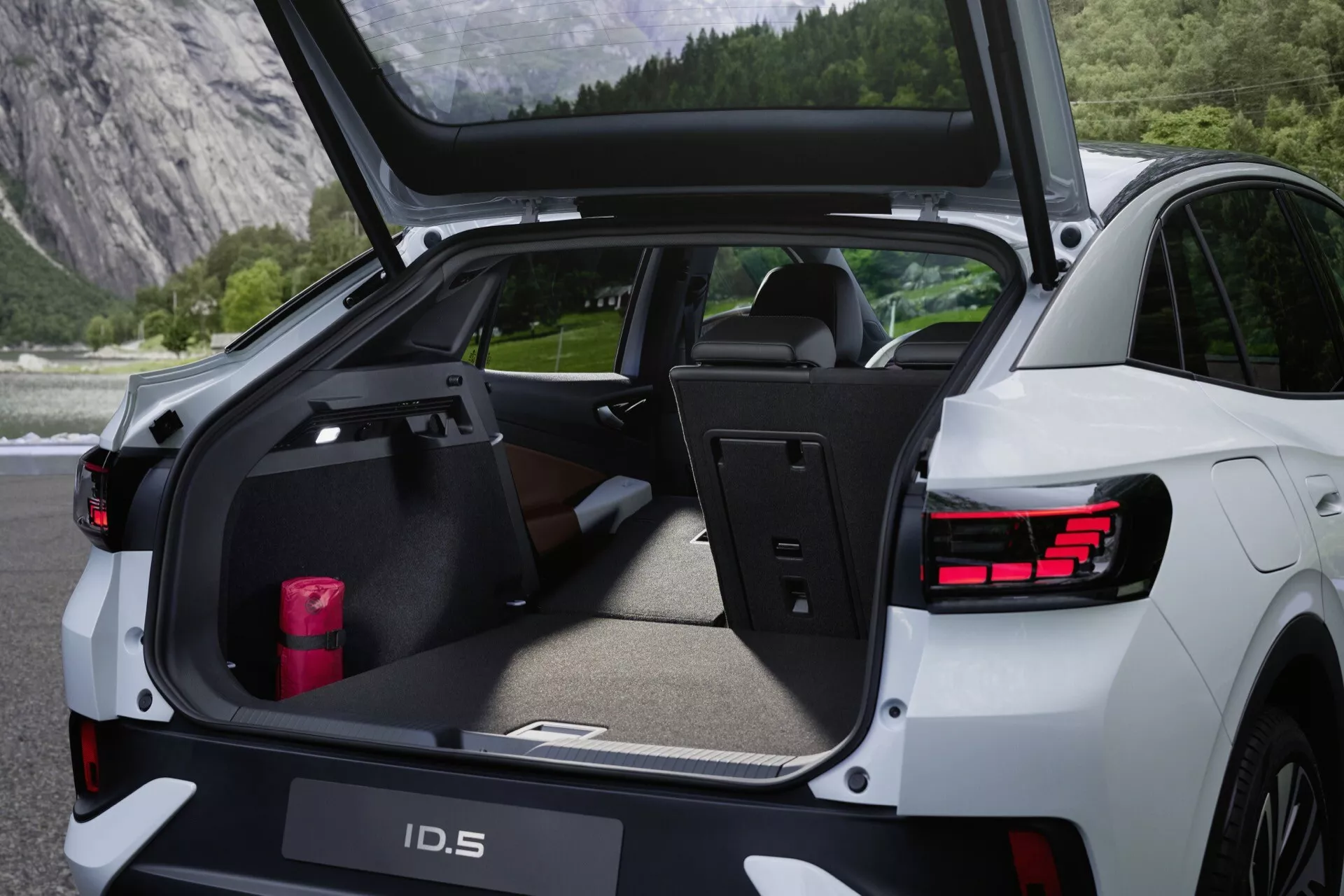 Zadný pohľad na Volkswagen ID.5 2022 s otvoreným zadným kufrom, ukazujúcim batožinový priestor, zdôrazňujúcim jeho praktickosť a kapacitu nákladu. Obrázok zvýrazňuje senzory na zadnom nárazníku, LED zadné svetlá, elektrické víko kufra, vysoko kvalitný interiér, zadné sedadlá s deleným sklopením, vysúvací kryt batožinového priestoru, plochý úložný priestor s ochrannou podložkou a lakťovú opierku s držiakmi na poháre. 'ID.5' je viditeľné na dvierkach kufra.