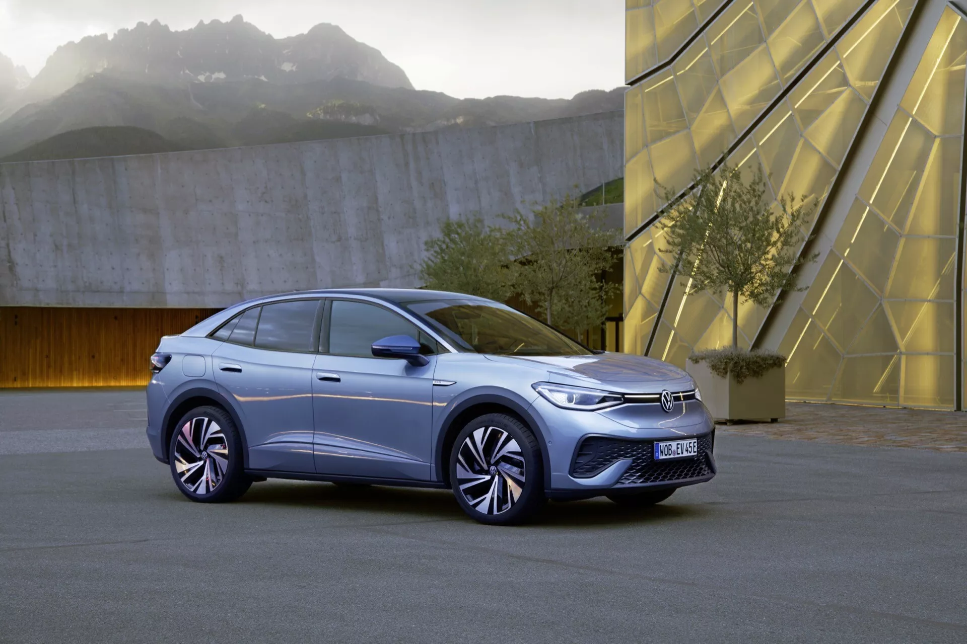 Trojštvrtinový pohľad spredu na prvú generáciu VW ID.5 2022, hatchback alebo crossover SUV. Obrázok zvýrazňuje ľavú prednú stranu vozidla, ukazuje LED svetlá, minimalistickú prednú mriežku, aerodynamický dizajn kolies a časť kapoty, blatníka a čelného skla. Mestské prostredie s geometrickými štruktúrami v pozadí dodáva moderný kontext. Senzory na prednom nárazníku naznačujú pokročilé asistenčné funkcie pre vodiča.