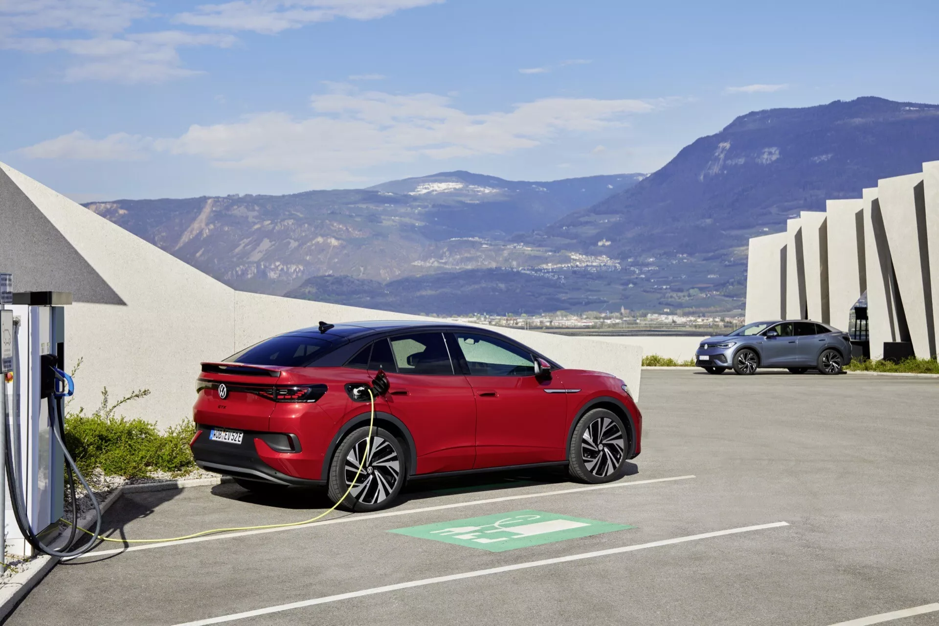 Obrázok ukazuje moderný červený hatchback VW ID.5 2022 zaparkovaný v nabíjacom boxe, čo naznačuje jeho elektrický alebo plug-in hybridný charakter. Vozidlo je zobrazené z profilu s jasným pohľadom na jeho elegantné línie karosérie, aerodynamický dizajn a otvorený nabíjací port pripojený k nabíjacej stanici viditeľným káblom. Kontext zahŕňa vyhradené zelené parkovacie miesto, abstraktné architektonické štruktúry, otvorené nebo a horský terén, čo zdôrazňuje ekologicky uvedomelé prostredie.