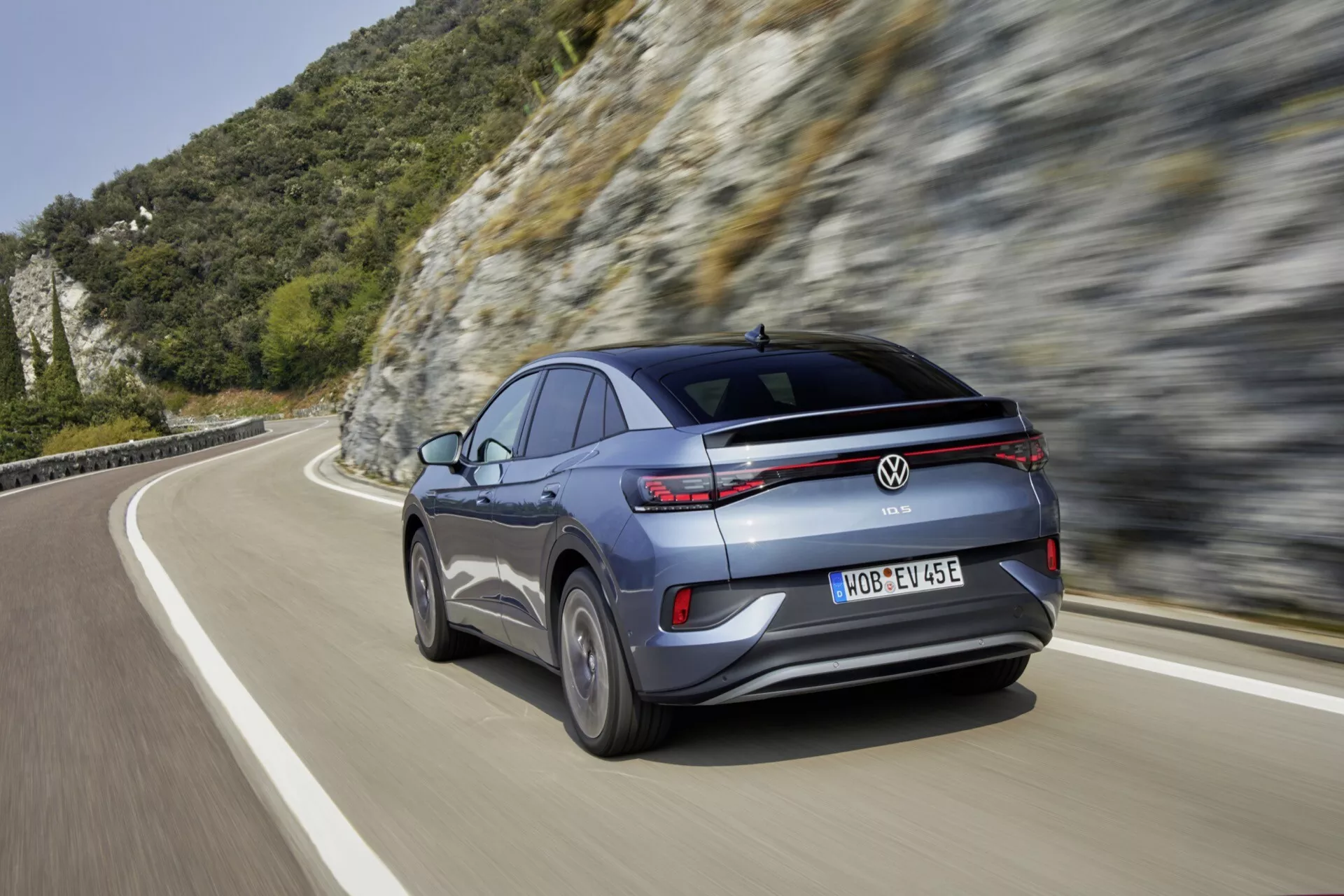 Moderné crossover SUV Volkswagen ID.5 2022 prvej generácie v pohybe, zobrazené zo zadného trojštvrťového uhla. Vozidlo je sivé alebo strieborné s kovovým leskom, má ostré, horizontálne LED zadné svetlá, viaclúčové zliatinové kolesá a bočné spätné zrkadlo v rovnakej farbe ako karoséria s integrovaným smerovým svetlom. Zadný nárazník obsahuje odrazky, senzory a zadnú kameru. Obrázok bol zachytený na horskej alebo kopcovitej ceste počas dňa a vyvoláva pocit rýchlosti a pokročilého automobilového výkonu.