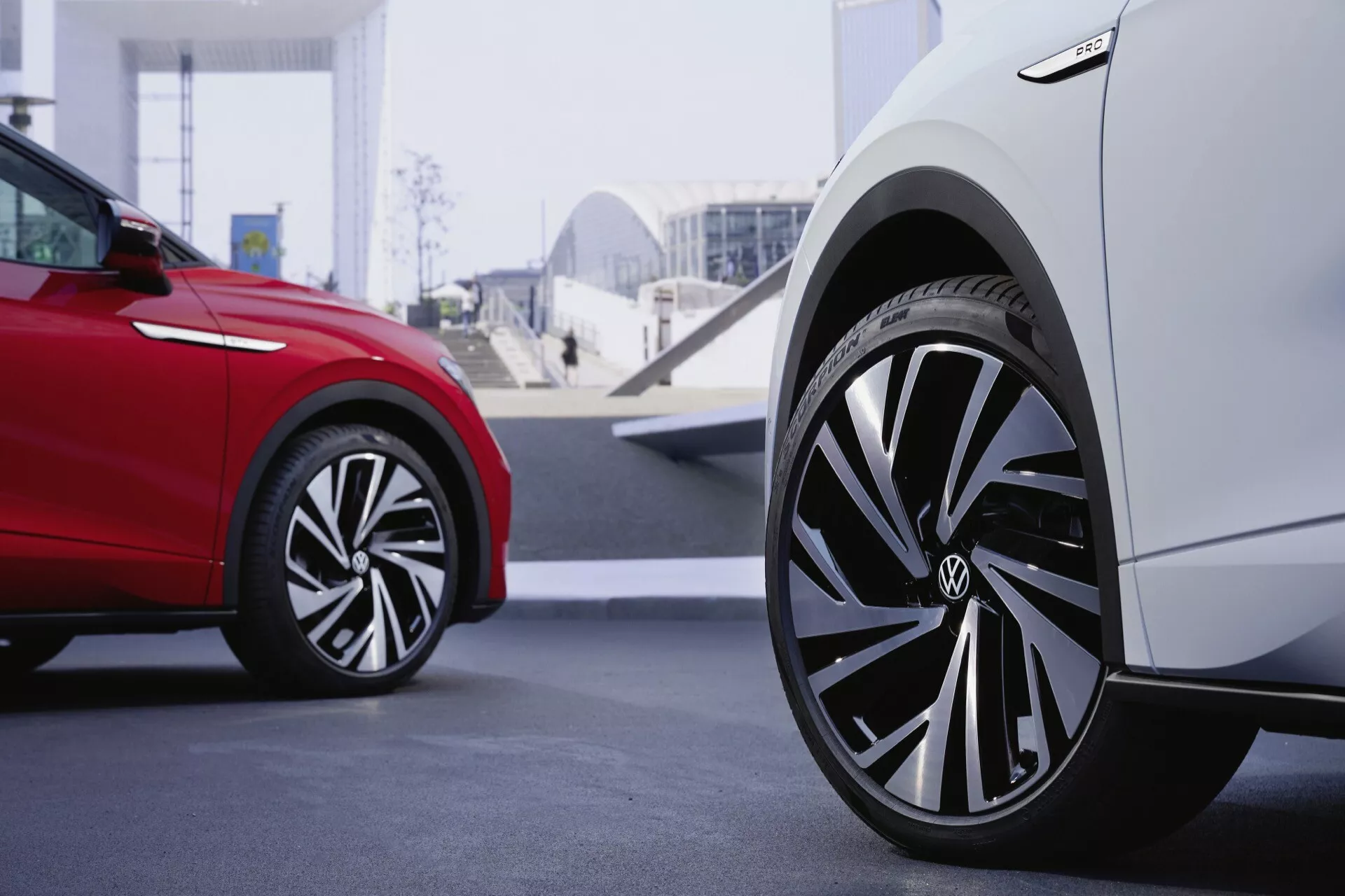 Predná časť bieleho Volkswagen ID.5 2022, zvýrazňujúca dizajn predného blatníka a kolesa s moderným aerodynamickým hliníkovým diskom. Hĺbka ostrosti sa zameriava na behúň, bočnicu a koleso bieleho auta, zatiaľ čo červené auto v pozadí vytvára bokeh efekt, zdôrazňujúc detailný pohľad na biele auto. Obrázok je urobený z nízkeho bočného uhla v modernom mestskom prostredí, prezentujúc sofistikovaný dizajn a dynamiku auta.