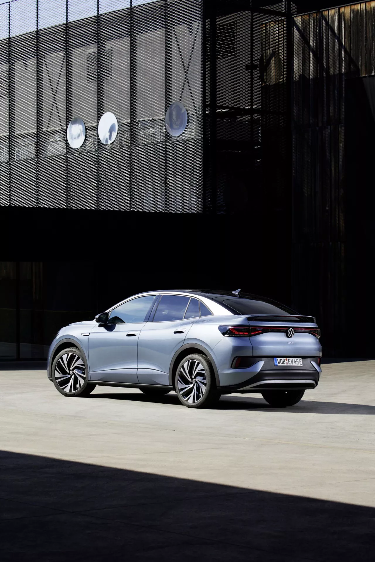 Obrázok sedanu VW ID.5 2022 (1. generácia) z pohľadu zozadu zo šikminy, ukazujúci jeho elegantný a moderný dizajn s veľkými hliníkovými kolesami, kovovým lakom, šikmou strechou v štýle kupé, jemným spoilerom na kufri, tenkými LED zadnými svetlami, tónovanými oknami a integrovanými výfukovými otvormi, zasadený do mestského prostredia s geometrickými architektonickými prvkami v pozadí.
