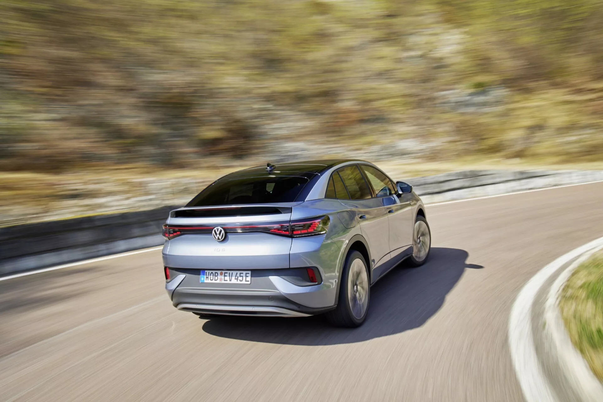 Zadný trojštvrťový pohľad na pohybujúce sa VW ID.5 2022 (1. generácia) za denného svetla, zachytený z nižšieho uhla kamery blízko zadného kolesa. Obrázok zdôrazňuje kupéovitú sklonenú líniu strechy, horizontálne zadné svetelné zoskupenia, centrálne umiestnené logo výrobcu pod integrovaným zadným spojlerom, centrálne umiestnenú registračnú značku, anténu v tvare plutvy žraloka a strešné okno/mesačné okno. Pohyb vozidla je naznačený rozmazaným pozadím a skreslením kolies, čo naznačuje strednú až vysokú rýchlosť, s reflexnými povrchmi farby a zadného skla, naznačujúcimi slnečný deň.