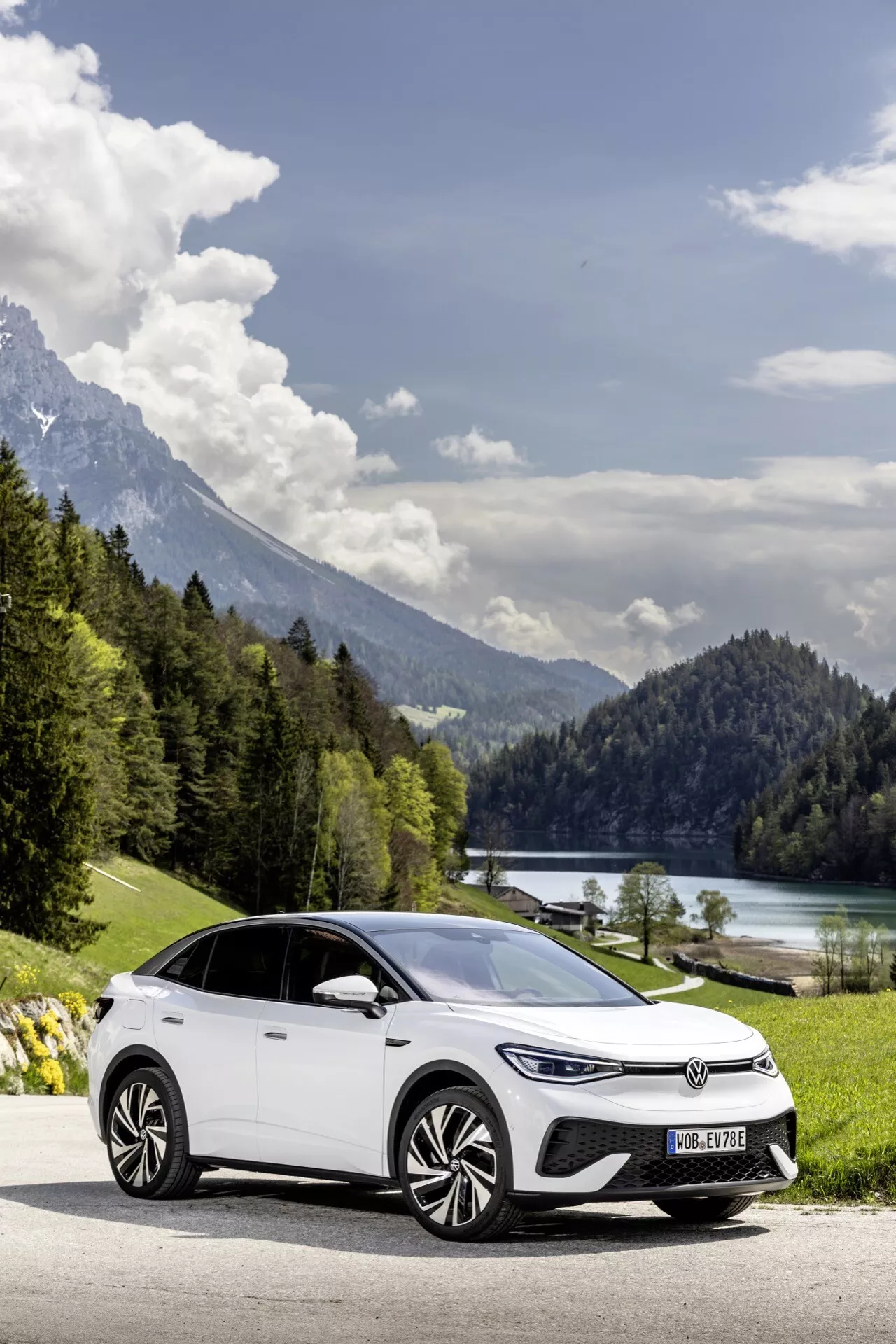 Obrázok moderného elektrického hatchbacku, VW ID.5 2022 (1. generácia), umiestneného na ceste s malebným pozadím jazera a hôr. Auto je zachytené z predného trojštvrťového uhla, zobrazujúceho jeho aerodynamické zliatinové kolesá, LED denné svetlá, dizajn svetlometu a hladké línie karosérie. Prirodzené osvetlenie a malebné prostredie zvýrazňujú ekologický aspekt vozidla s dostatočným priestorom okolo pre pocit otvorenosti.