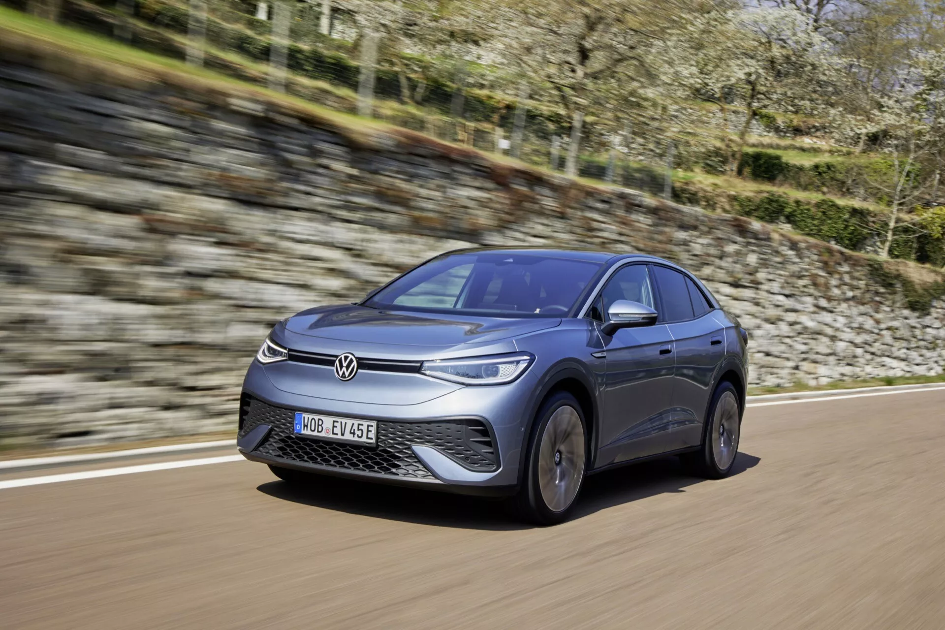 Dynamický pohľad na Volkswagen ID.5 z roku 2022 (prvá generácia) zachytený z nízkeho, predného trojštvrťového uhla, zobrazujúci predný nárazník, mriežku, kapotu, čelné sklo a profil na strane vodiča. Auto, natreté v metalickej farbe, sa pohybuje po dláždenej ceste, čím predvádza svoj elegantný, aerodynamický dizajn a technológiu elektrického vozidla. Predná časť, zliatinové kolesá a značka výrobcu sú jasne viditeľné, zdôrazňujúc pokročilú osvetľovaciu technológiu a uzavretú masku chladiča typickú pre elektrické vozidlá.