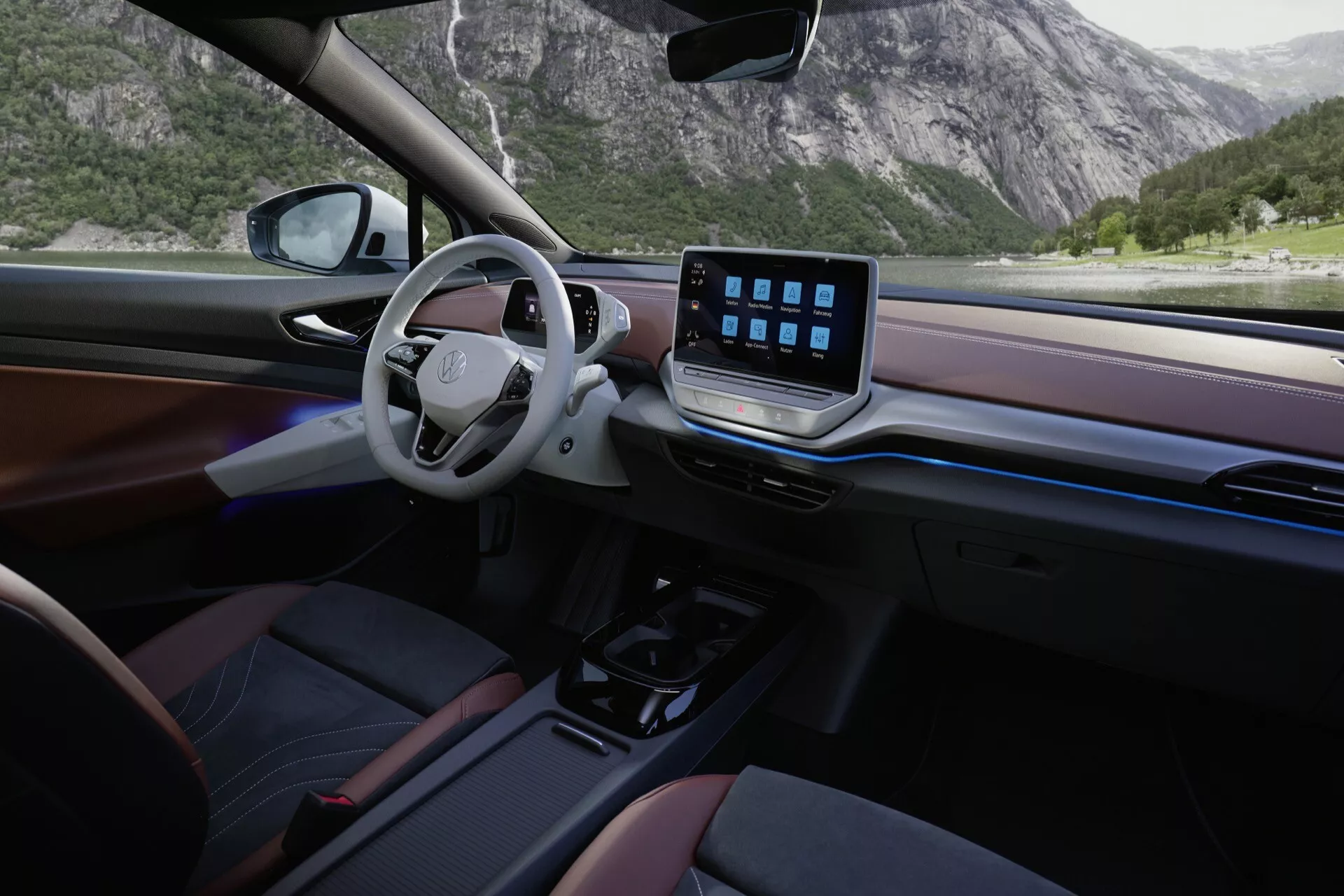 Interiér VW ID.5 2022. Uhol kamery poskytuje pohľad zo zadného pravého sedadla spolujazdca, ukazujúcý elegantný minimalistický dizajn a luxusný dojem. Vľavo umiestnený volant nemá tradičný prístrojový panel, čo naznačuje prítomnosť digitálneho displeja. V strede palubnej dosky je výrazný dotykový displej, zdôrazňujúci pokročilé funkcie infotainment systému. Minimálne fyzické ovládacie prvky a automatická prevodovka naznačujú moderné dizajnové preferencie. Palubná doska a dverové panely obsahujú materiály s drevím efektom a tmavé plasty, čím zvyšujú prémiovú estetiku. Prirodzené svetlo prichádzajúce cez veľké bočné okno osvetľuje interiér, ktorý má tmavé čalúnenie s červenými prvkami.