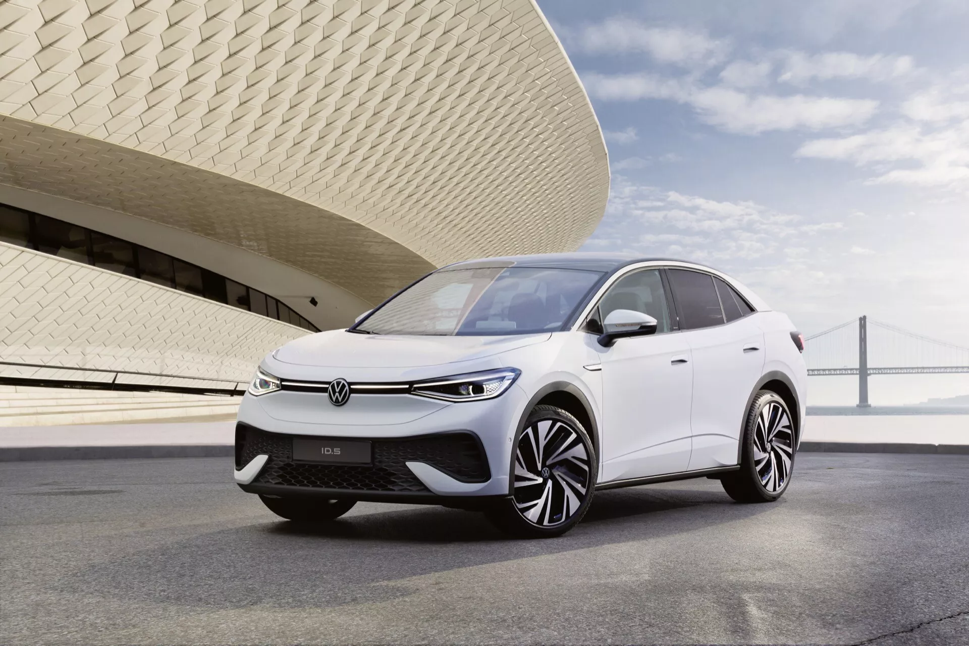 Obrázok Volkswagen ID.5 z roku 2022 (prvá generácia) v bielej farbe, umiestnený v trojštvťovom uhle, aby vynikol predok a bok vodiča. Pohľad zahŕňa prednú mriežku, elegantné LED svetlomety, aerodynamickú kapotu a zložité liatinové kolesá. Zvýšený postoj auta a uzavretá mriežka naznačujú, že ide o elektrické vozidlo (EV) s pokročilými funkciami, pravdepodobne vrátane senzorov pre asistenčné technológie riadenia. Auto je zobrazené za denného svetla, zaparkované na hladkom chodníku s modernou budovou v pozadí, čo zvýrazňuje jeho súčasný a efektívny dizajn.