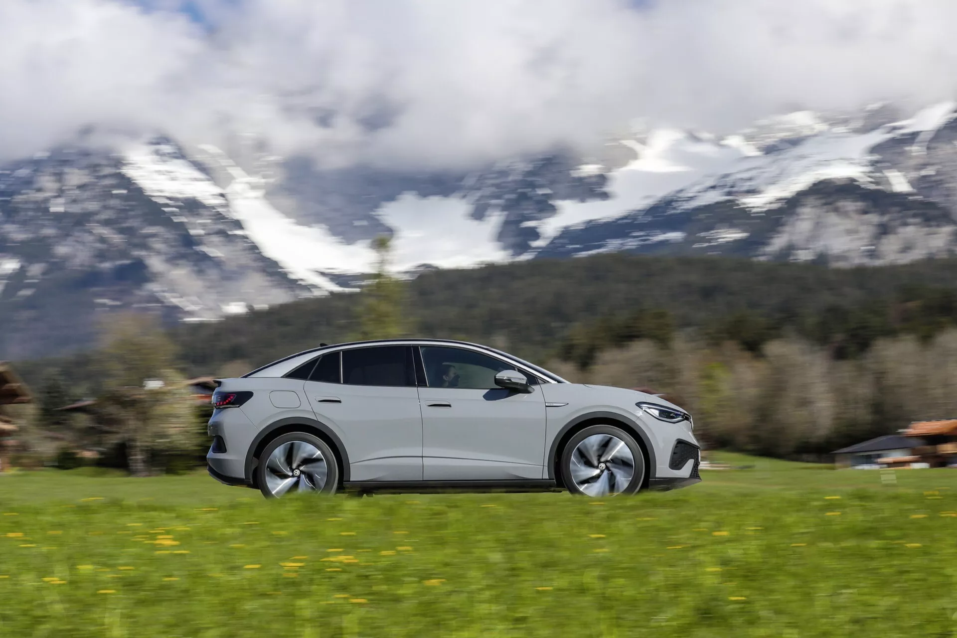 Dynamický záber moderného VW ID5 2022 (prvá generácia) v pohybe, zachytený technikou panorámovania, ktorá zobrazuje rýchlosť a pohyb. Pohľad z bočného profilu s aerodynamickými líniami, nízkoprofilovými pneumatikami a moderným dizajnom kolies. Vozidlo, pravdepodobne športový sedan s kupé siluetou a veľkými oknami, prípadne vrátane panoramatickej strechy, je ostré, keď sa pohybuje zľava doprava proti rozmazanému pozadiu. S premalovanými kľučkami dverí a bočnými zrkadlami. Scenéria naznačuje vidiecke či malebné prostredie s trávnatým terénom a horami v pozadí.