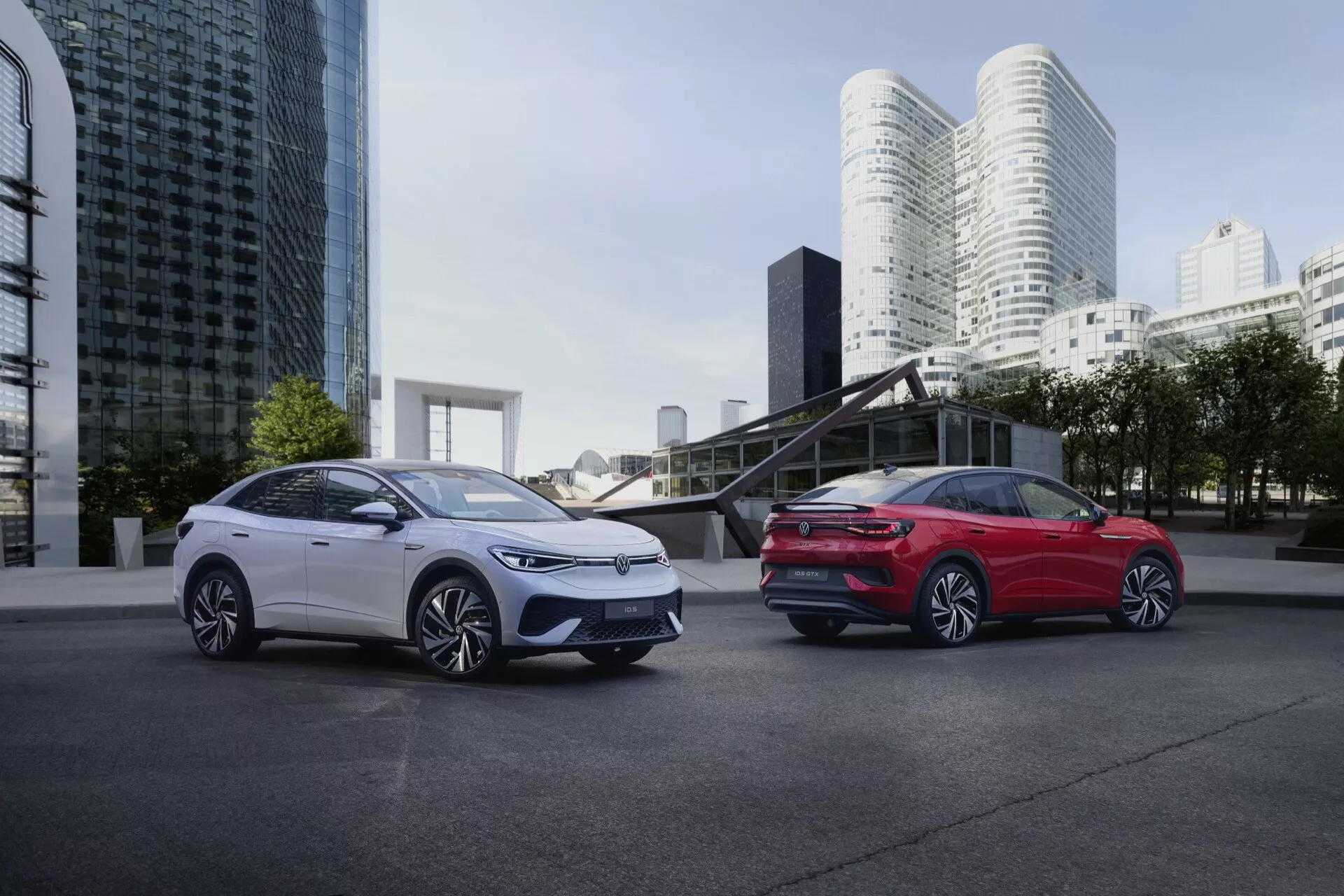 Dve elegantné a moderné elektrické alebo hybridné vozidlá VW ID.5 2022 (1. generácia) na pozadí s modernou architektúrou. Obrázok, urobený z nízkeho uhla, zdôrazňuje rozmery vozidiel. Biely voz naľavo ukazuje svoju ľavú prednú stranu, vrátane mriežky, dizajnu svetlometov, kontúr kapoty a kolesa na strane vodiča. Červený voz napravo zobrazuje svoju pravú zadnú stranu s viditeľným dizajnom zadných svetiel, zadného nárazníka a krivky zadnej štvrtiny panelu. Obe vozidlá majú aerodynamické kolesá z ľahkých zliatin so zložitými vzormi lúčov a ich lesklý povrch karosérie odráža svetlo okolia skoro ráno alebo neskoro popoludní.