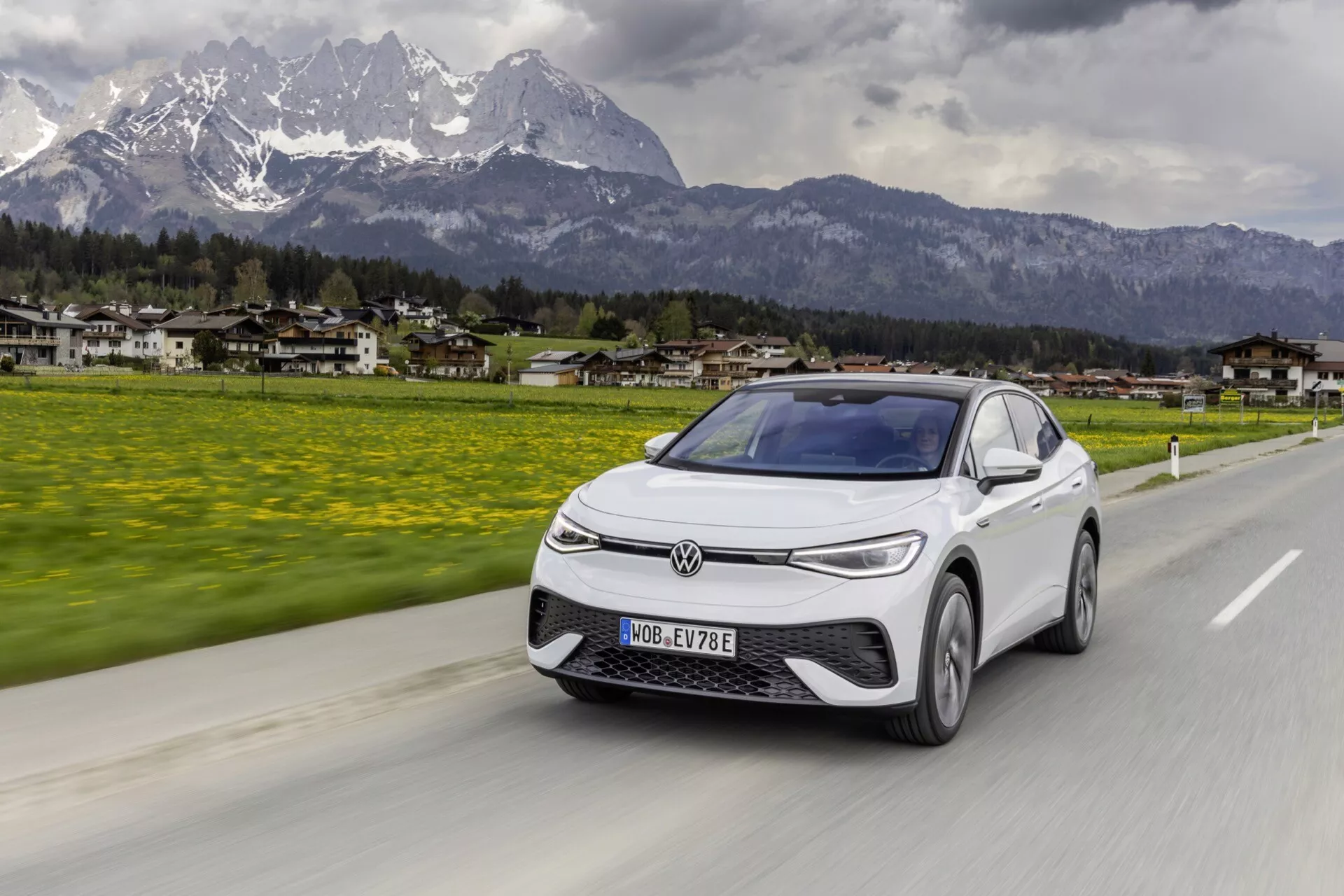 Obrázok zobrazuje auto VW ID.5 2022 prvej generácie jazdiace po ceste s malebným pozadím hôr a malého mesta. Fotené z trojštvrťového predného uhla, zameriava sa na ľavú prednú stranu, ukazuje mriežku, svetlomety, kapotu, čelné sklo a časť dverí vodiča a okna. Elegantné, aerodynamické elektrické vozidlo má uzavretú mriežku, úzke hranaté LED svetlomety, pokročilé aerodynamické línie a moderné disky na pohybujúcich sa kolesách. Auto je zobrazené v pohybe, s integrovanými indikátormi na bočných zrkadlách a panoramatickým čelným sklom, ktoré sa tiahne smerom k streche, čo naznačuje zlepšenú viditeľnosť vodiča.
